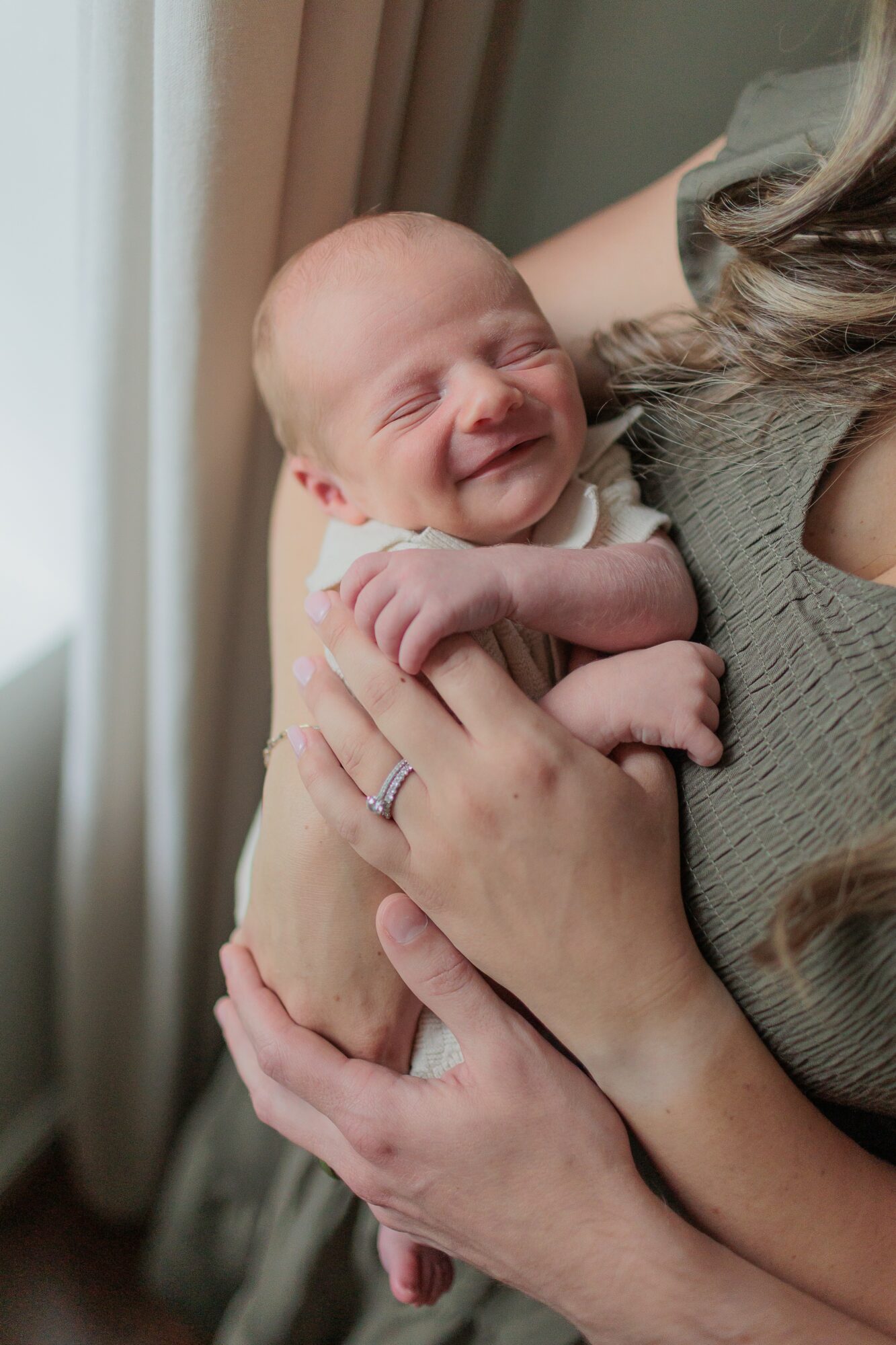 A smiling newborn baby being held by a woman, with natural light coming from a window on the left.