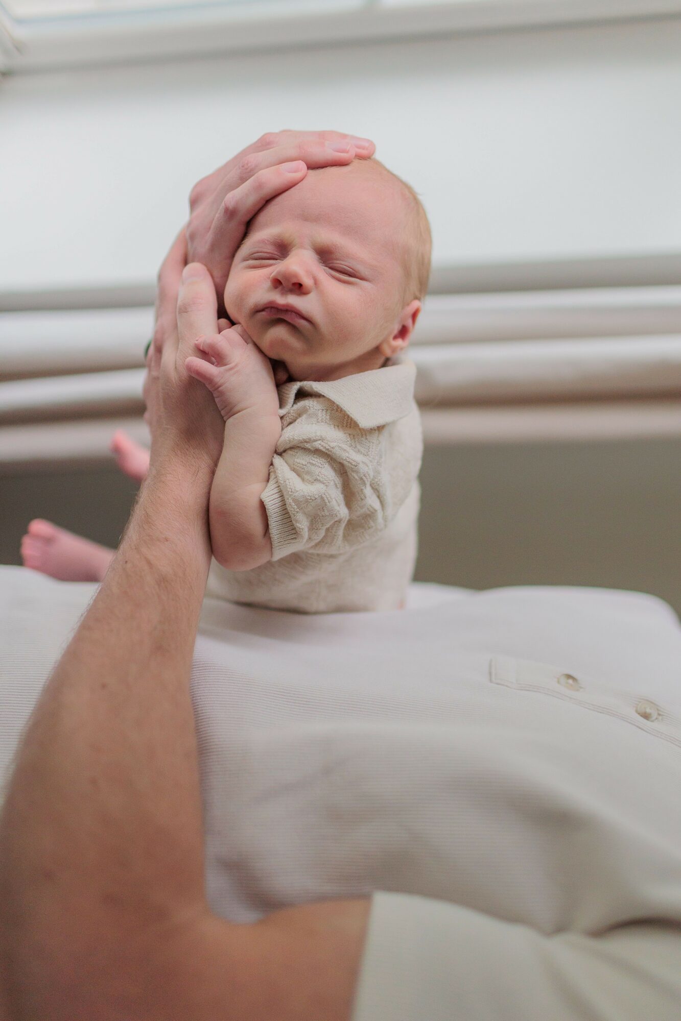 Baby with closed eyes resting head on adult's hand, adult's arm visible, in a hospital setting.