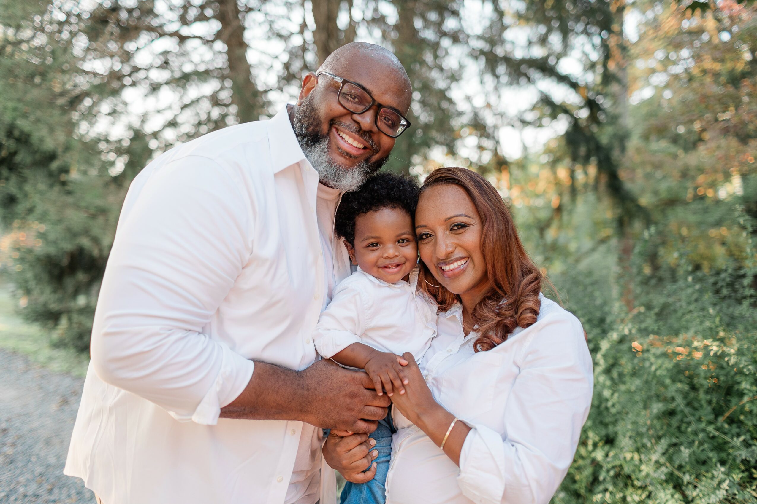 Smiling family of three outdoors, man, woman, and child, with trees in background.