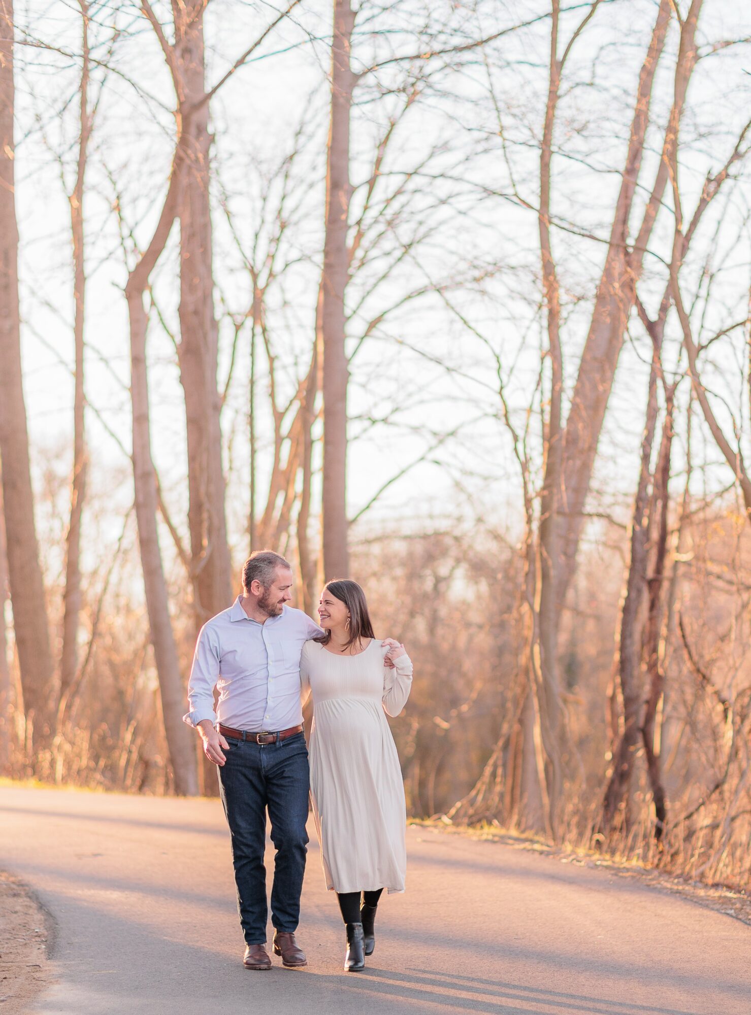 A couple walking on a road surrounded by leafless trees during daytime, holding hands and looking at each other.