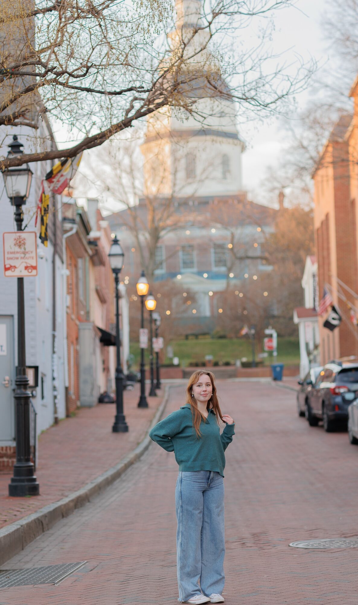 Young woman standing on a brick street with buildings and a church in the background, trees and street lamps visible.