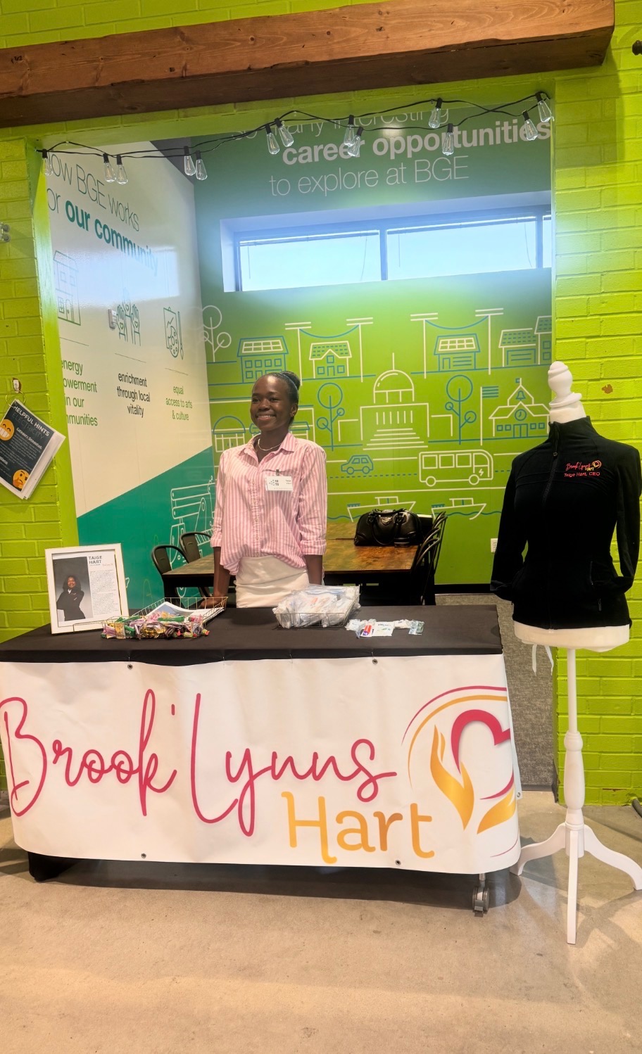 Person standing behind a table with a sign reading 'Brooklyn Lynus Hart' in a room with green walls and a window.