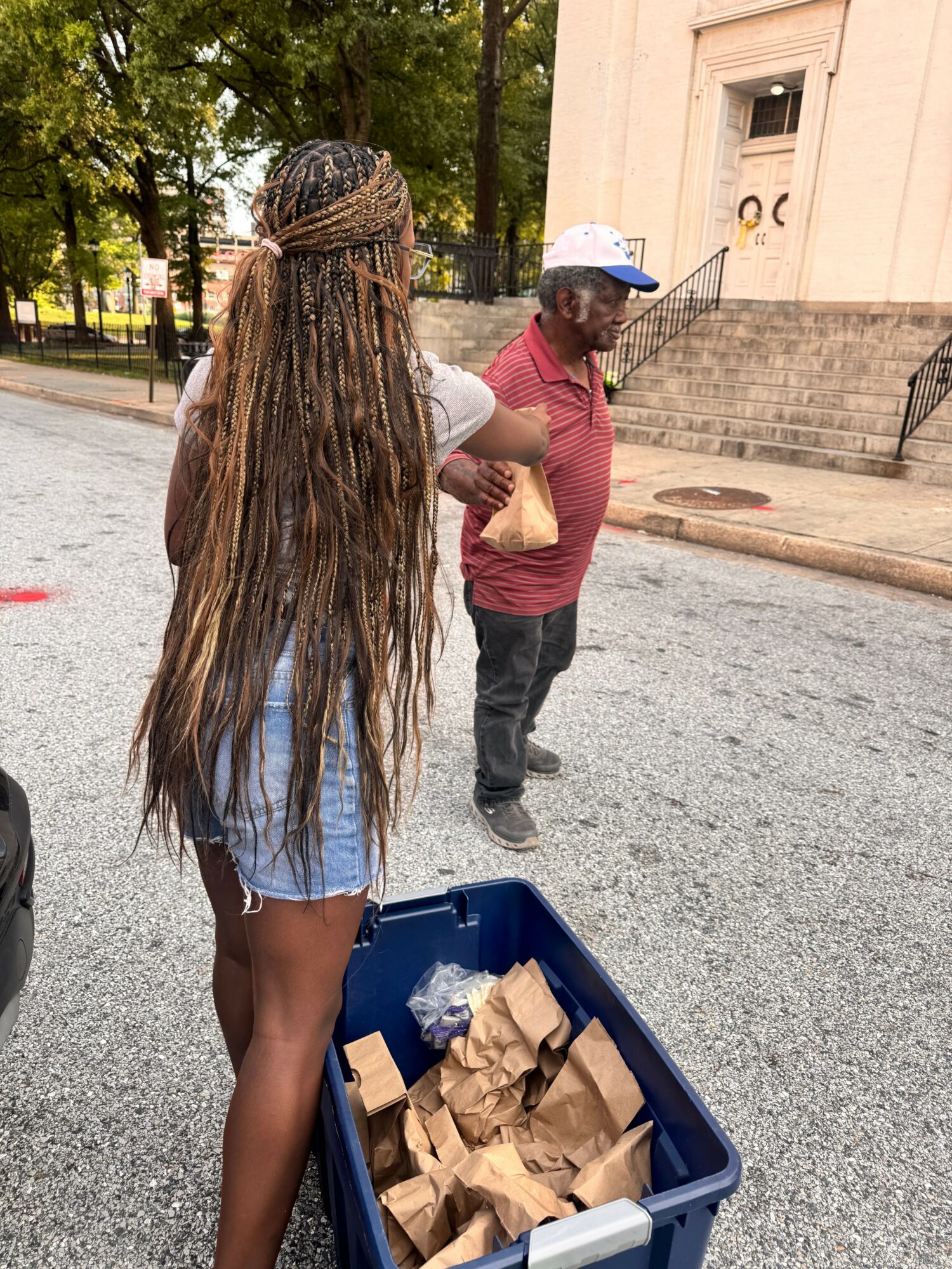 Person with long braided hair wearing a blue dress and white hat, standing next to a blue cart filled with paper bags, outdoors.