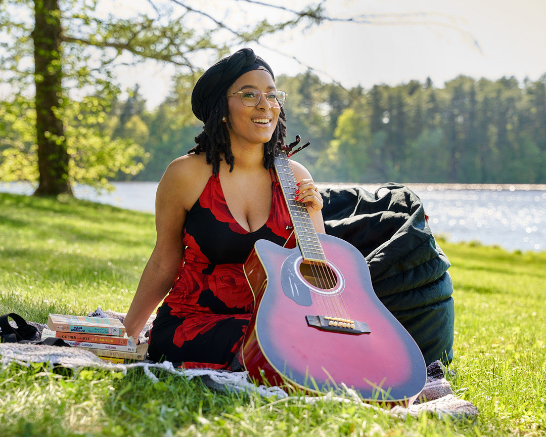 Woman sitting on grass with guitar, books, and a lake in background, smiling, outdoors on sunny day.