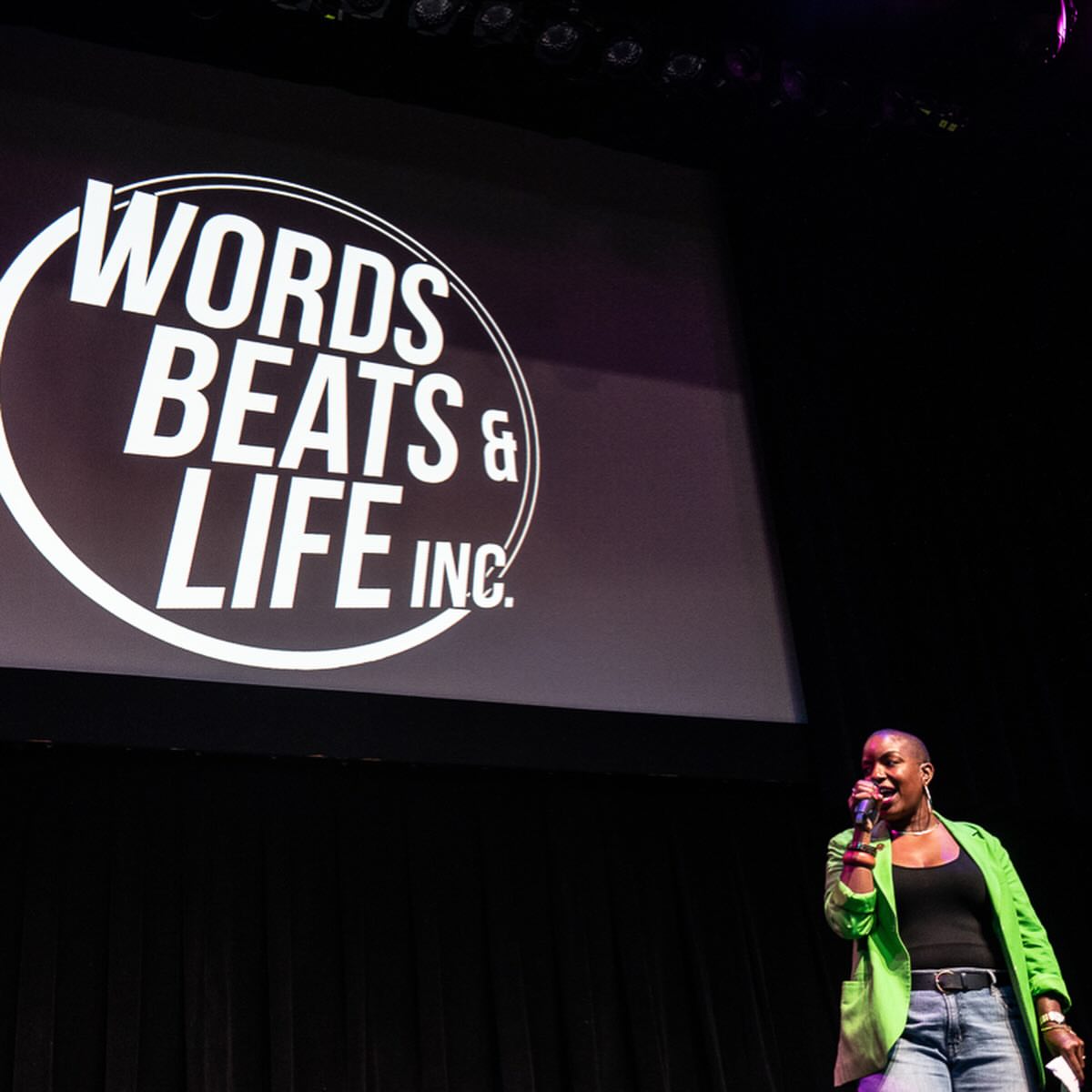 Person speaking on stage with a large screen displaying 'Words Beats & Life Inc.' behind them.