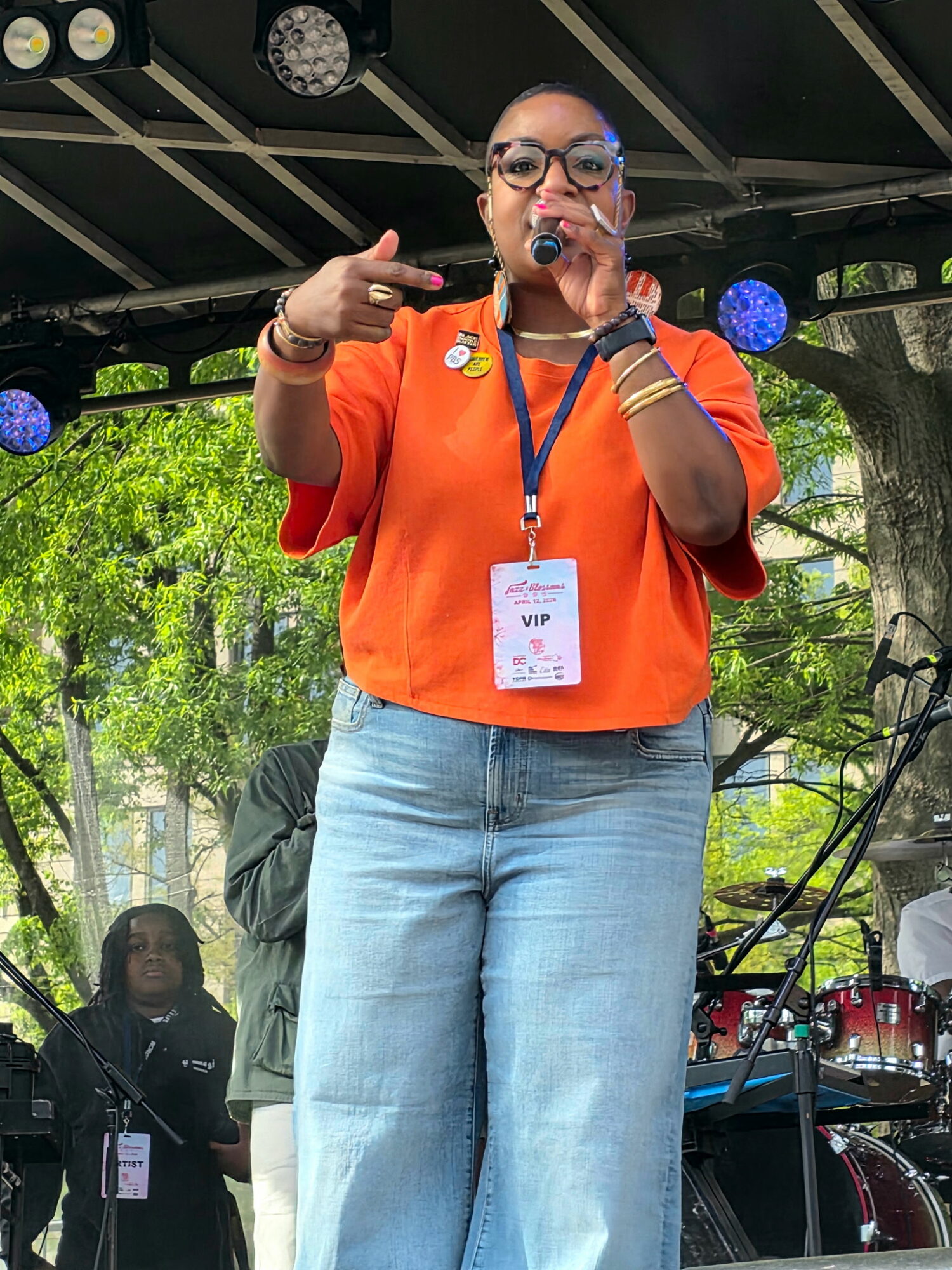 Woman speaking into microphone on stage, wearing orange top, jeans, glasses, outdoors with trees and stage equipment