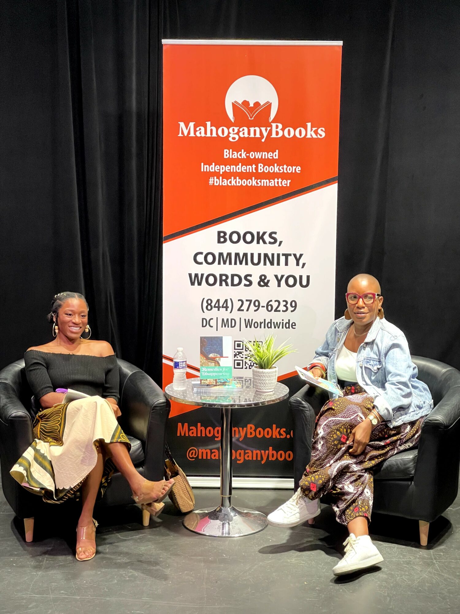 Two people sitting on black chairs with a table between them, in front of a MahoganyBooks banner, on a black stage.