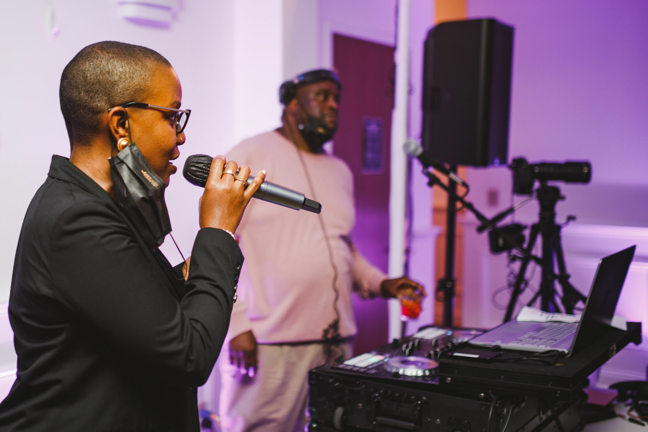 Person speaking into a microphone at an event with audio equipment and a person in the background.