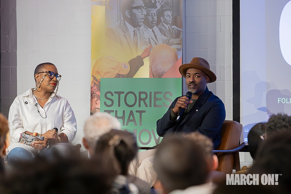 Two people sit on stage during a panel discussion, with an audience in front. One person holds a microphone, wearing a hat.