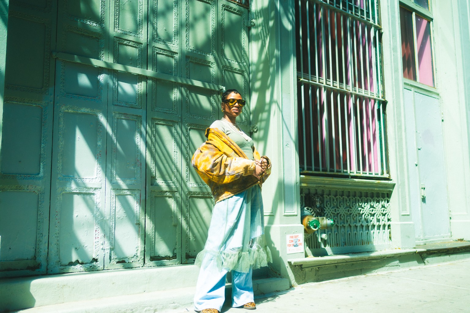 Person standing against a green wall with shadows, wearing sunglasses, a yellow jacket, and light-colored pants.