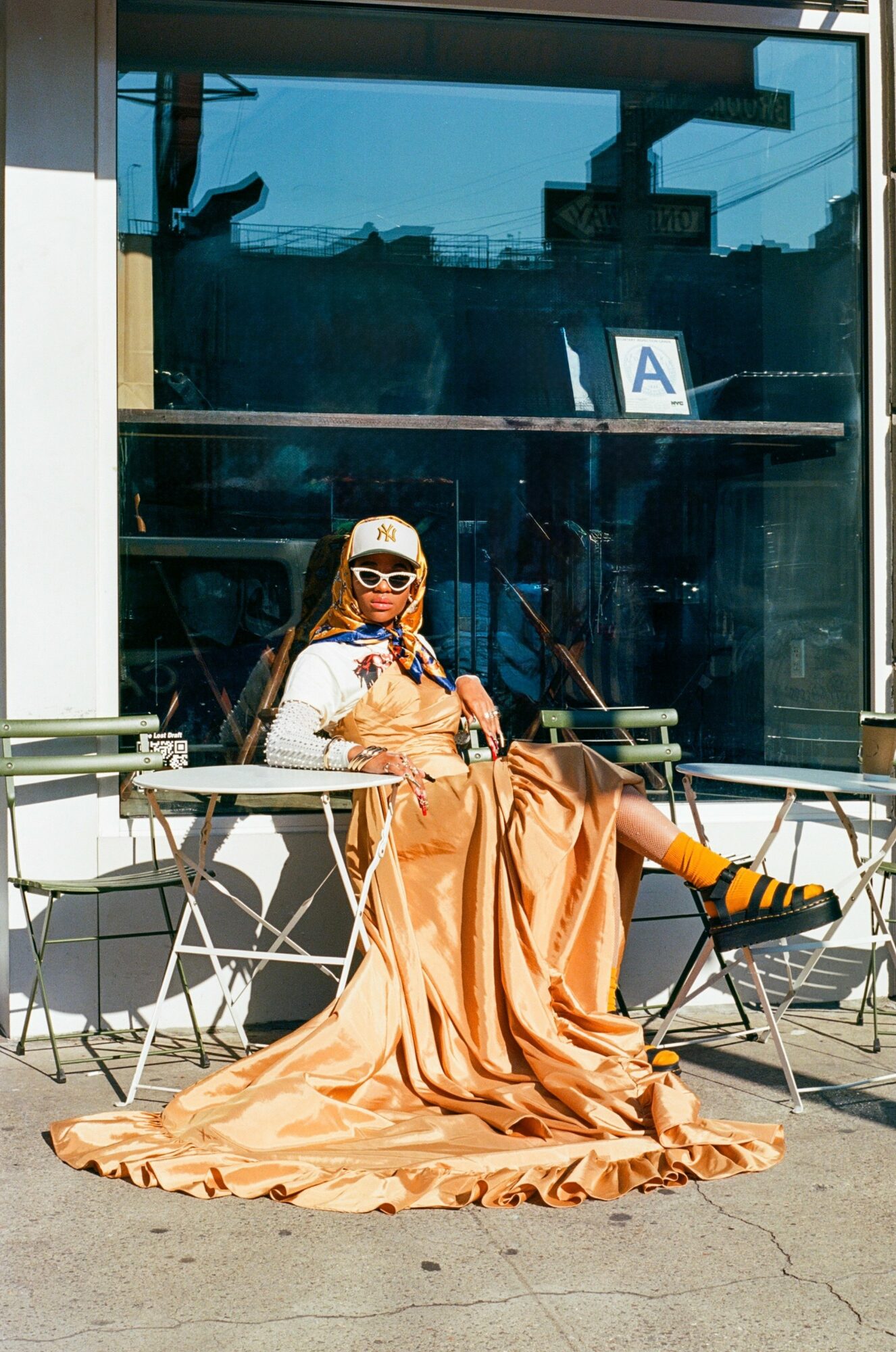 Person sitting outside a glass building, wearing sunglasses, a long dress, and sneakers, with a sign labeled 'A' behind them.