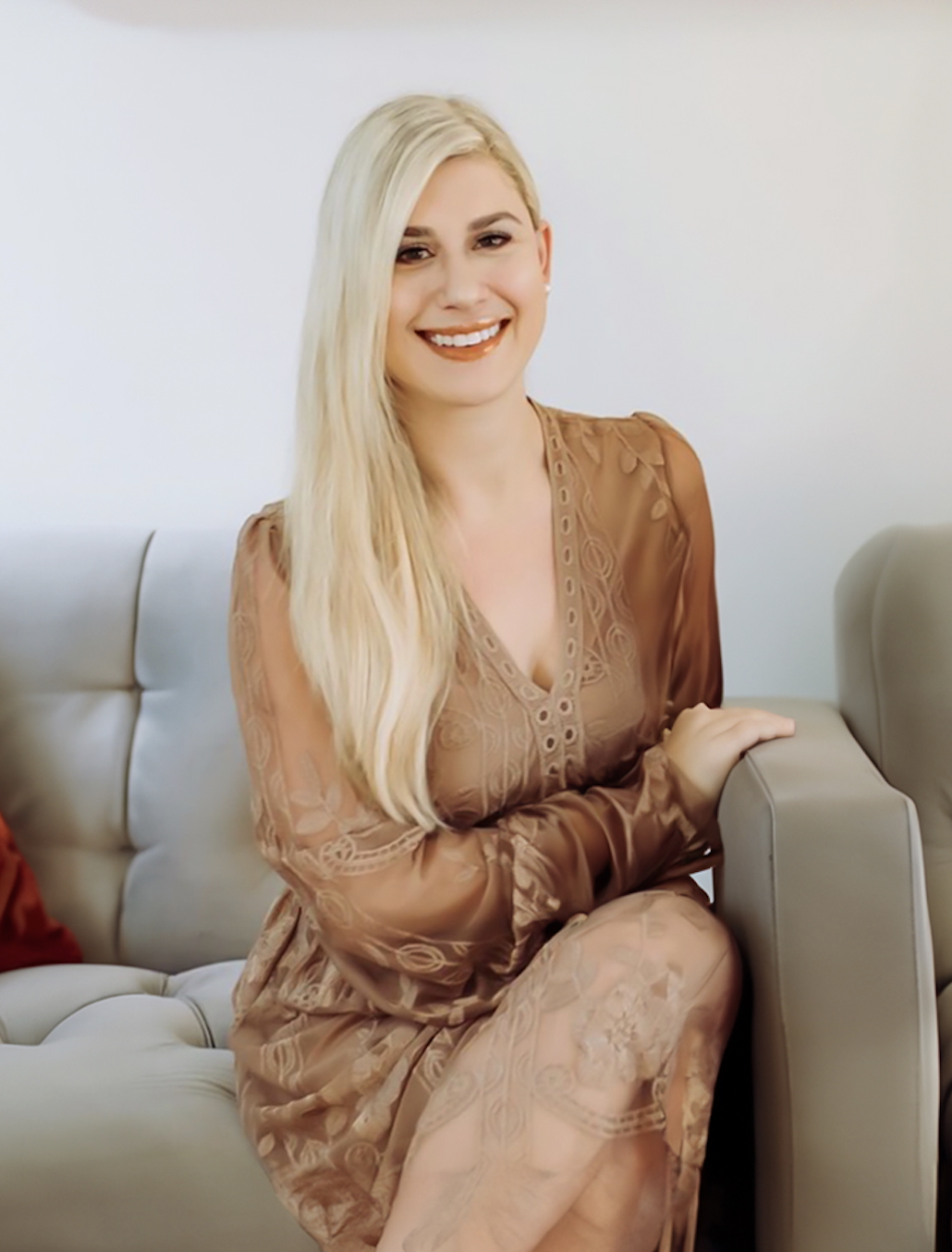 Young woman with long blonde hair smiling, sitting on a light-colored sofa, wearing a brown dress, in a bright room.