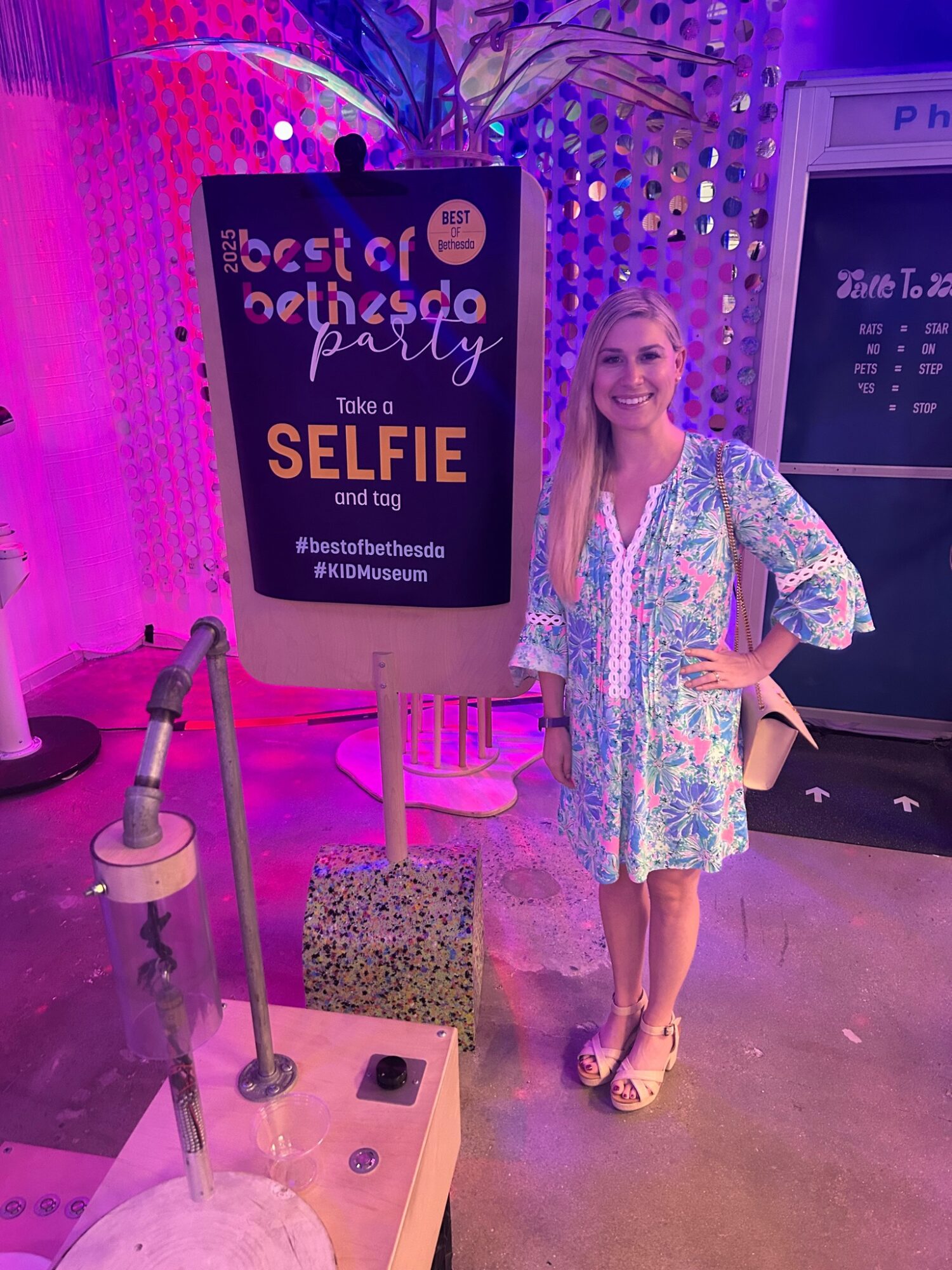 Smiling woman in a colorful dress taking a selfie at a party with a sign behind her, purple lighting, and a digital display.