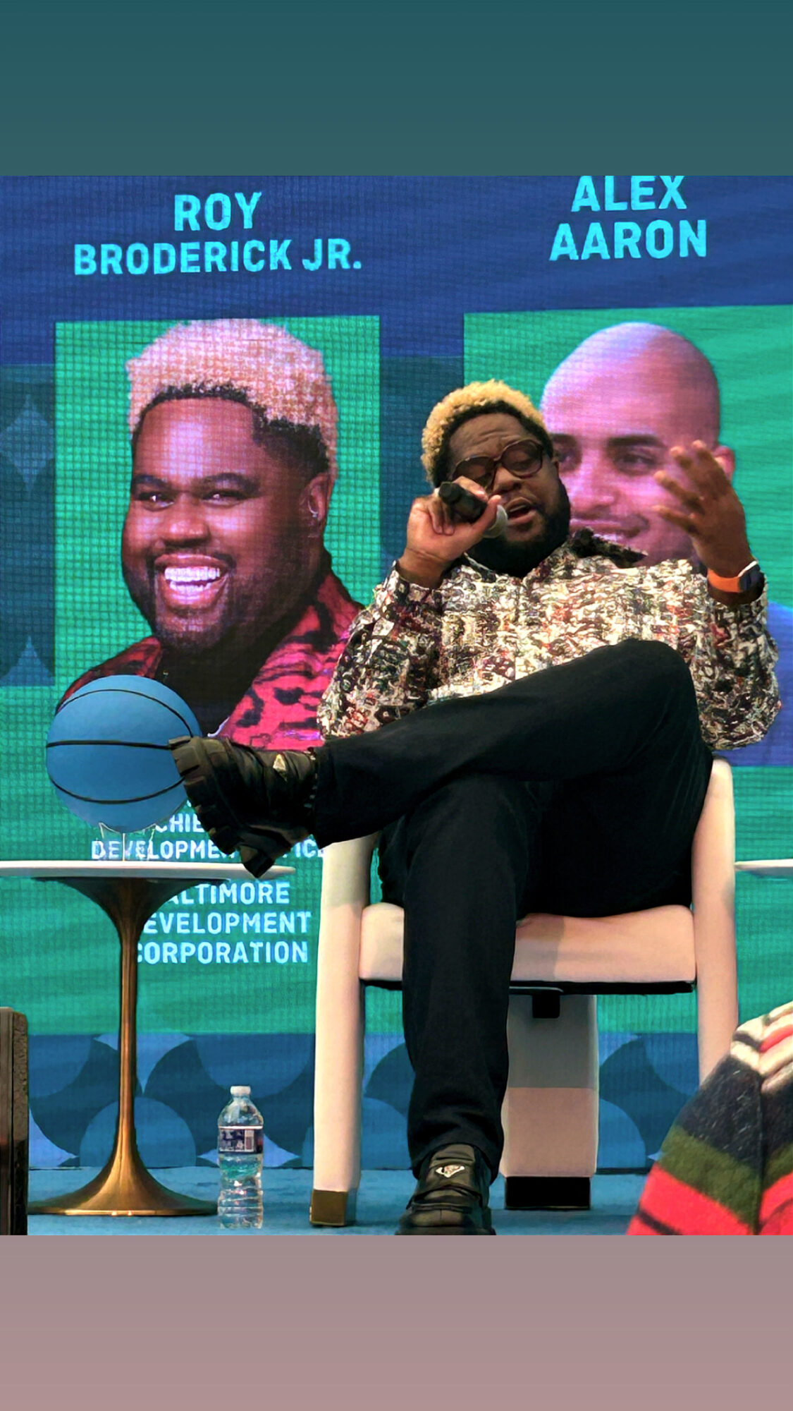 Two men sitting on chairs, one holding a microphone, with a colorful background and text, including a basketball and water bottle.