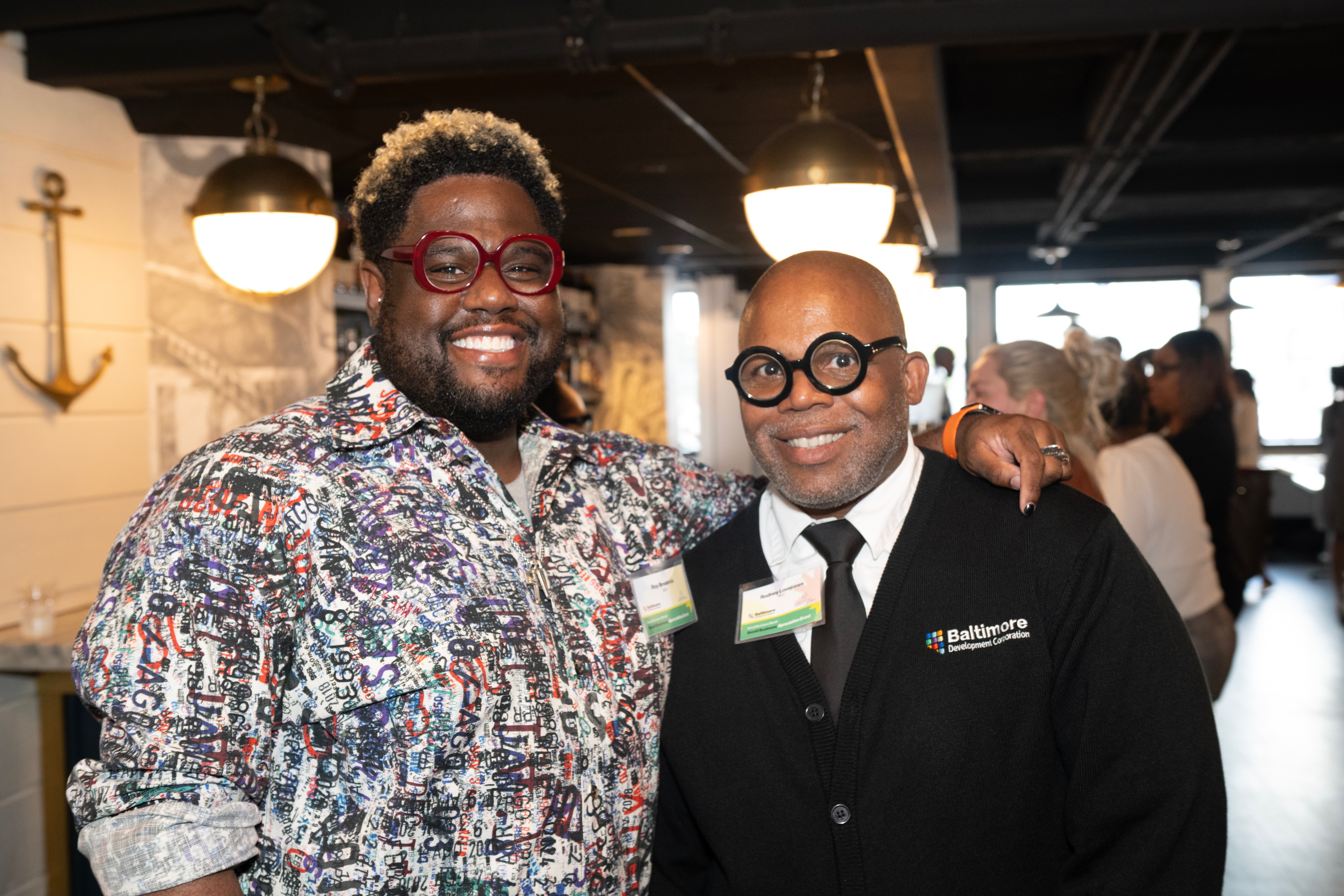 Two men smiling at an indoor event, one wearing a colorful patterned shirt and red glasses, the other in a black suit with glasses.