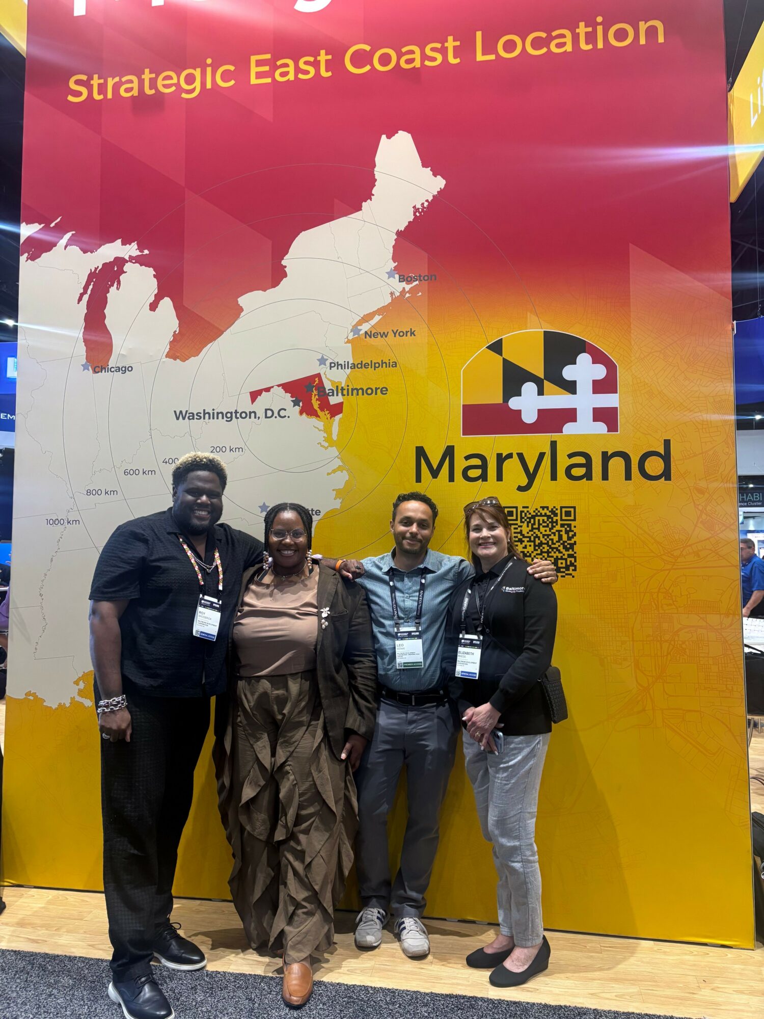 Three people standing in front of a Maryland tourism display with a map of Maryland and the state flag. The display reads 'Strategic East Coast Location.'