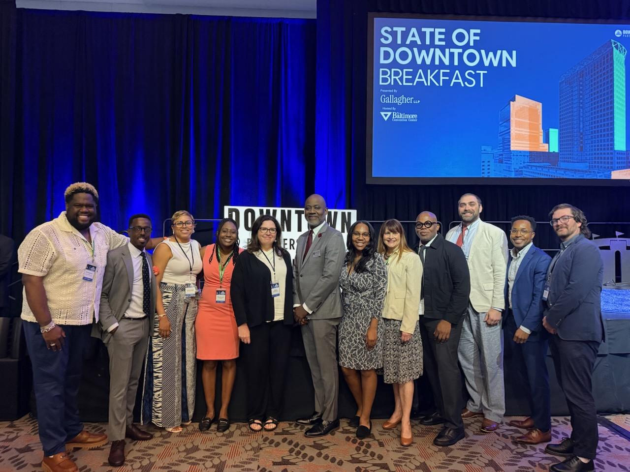 Group of people standing on stage at a breakfast event, with a screen displaying 'State of Downtown Breakfast' in the background.