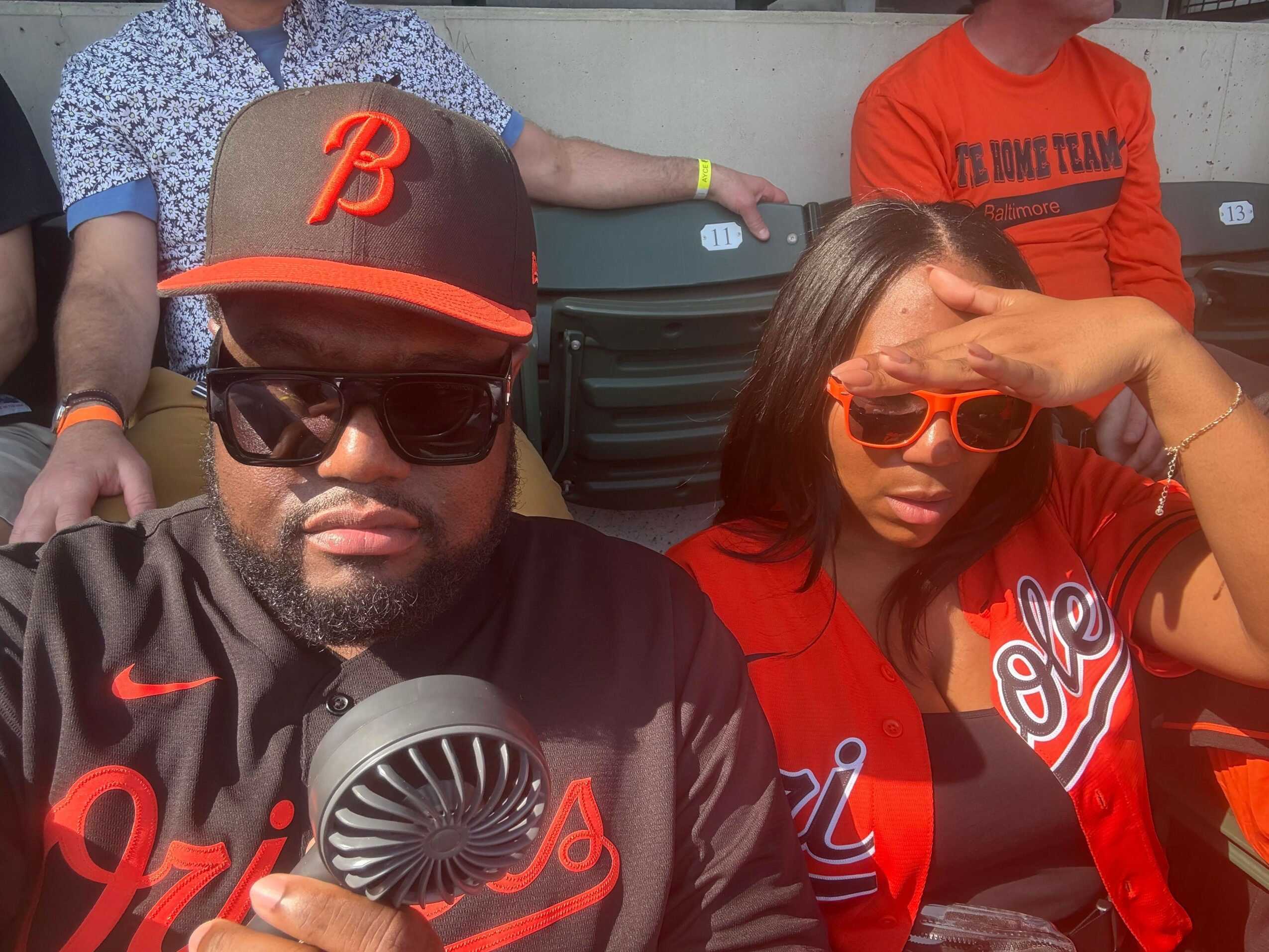 Two people wearing sunglasses and hats, one in a red shirt and the other in a patterned shirt, pose outdoors.