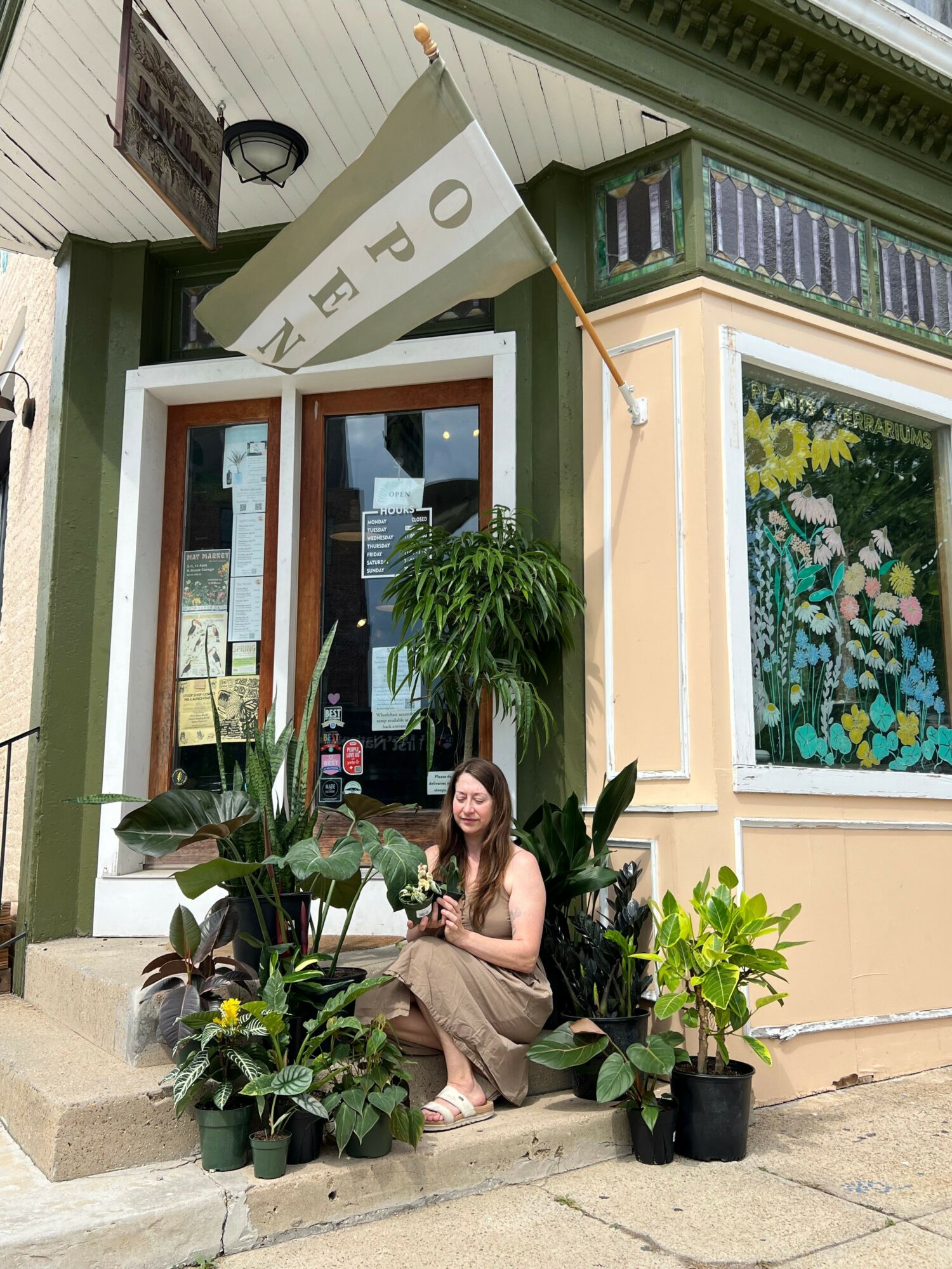 Woman sitting on sidewalk surrounded by potted plants outside a shop with an open sign, window display, and colorful artwork.