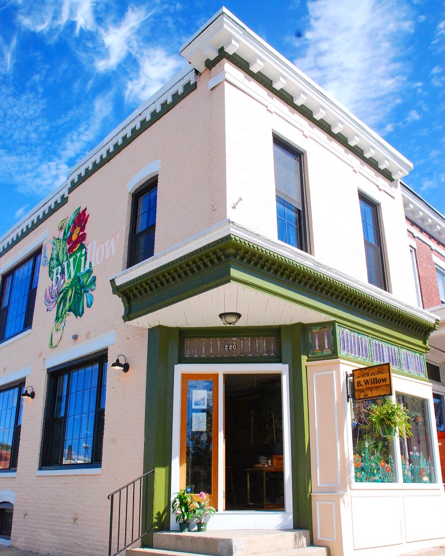 Corner of a two-story building with a green and white storefront, flower decoration, and a sign, against a blue sky.
