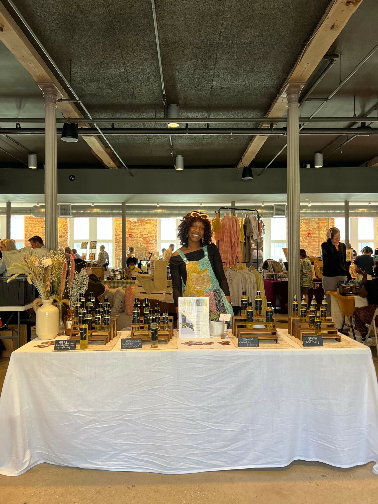 People browsing at a market stall with bottles and products, inside a building with large windows and natural light.