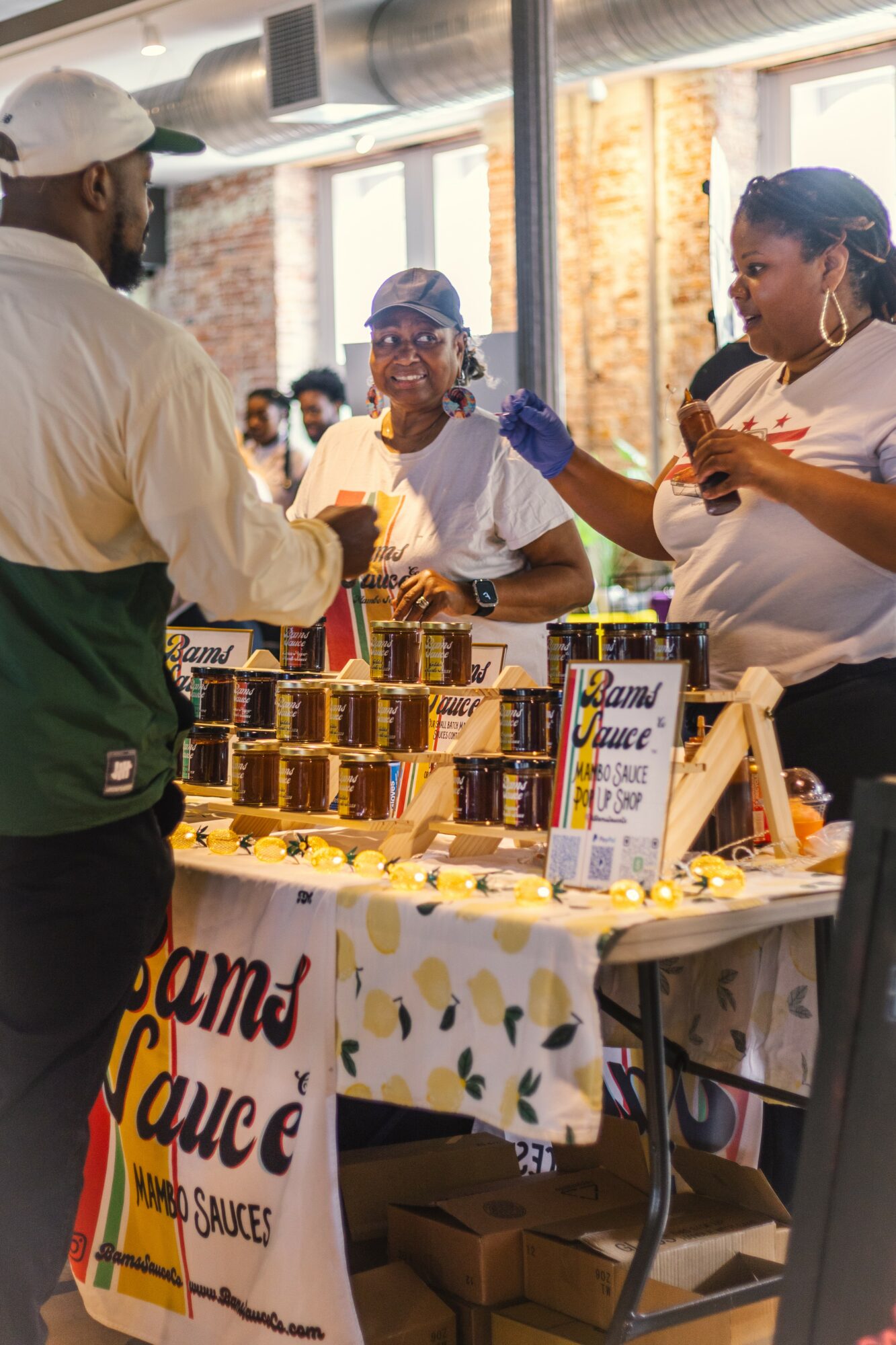 Three women behind a table selling jars of sauce at an indoor market, with a man in front of them. Bright natural light.