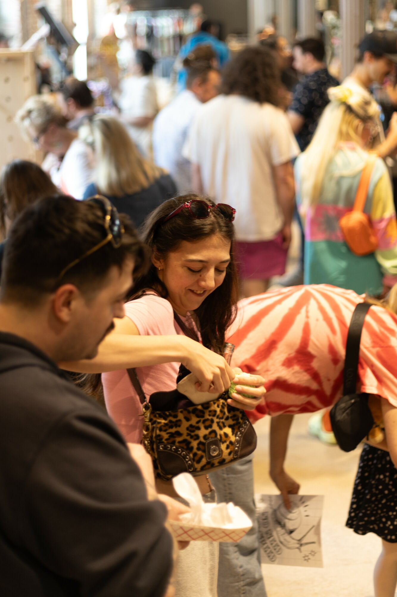 People gathered outdoors, some smiling and interacting, with a woman holding a leopard-print bag in focus.