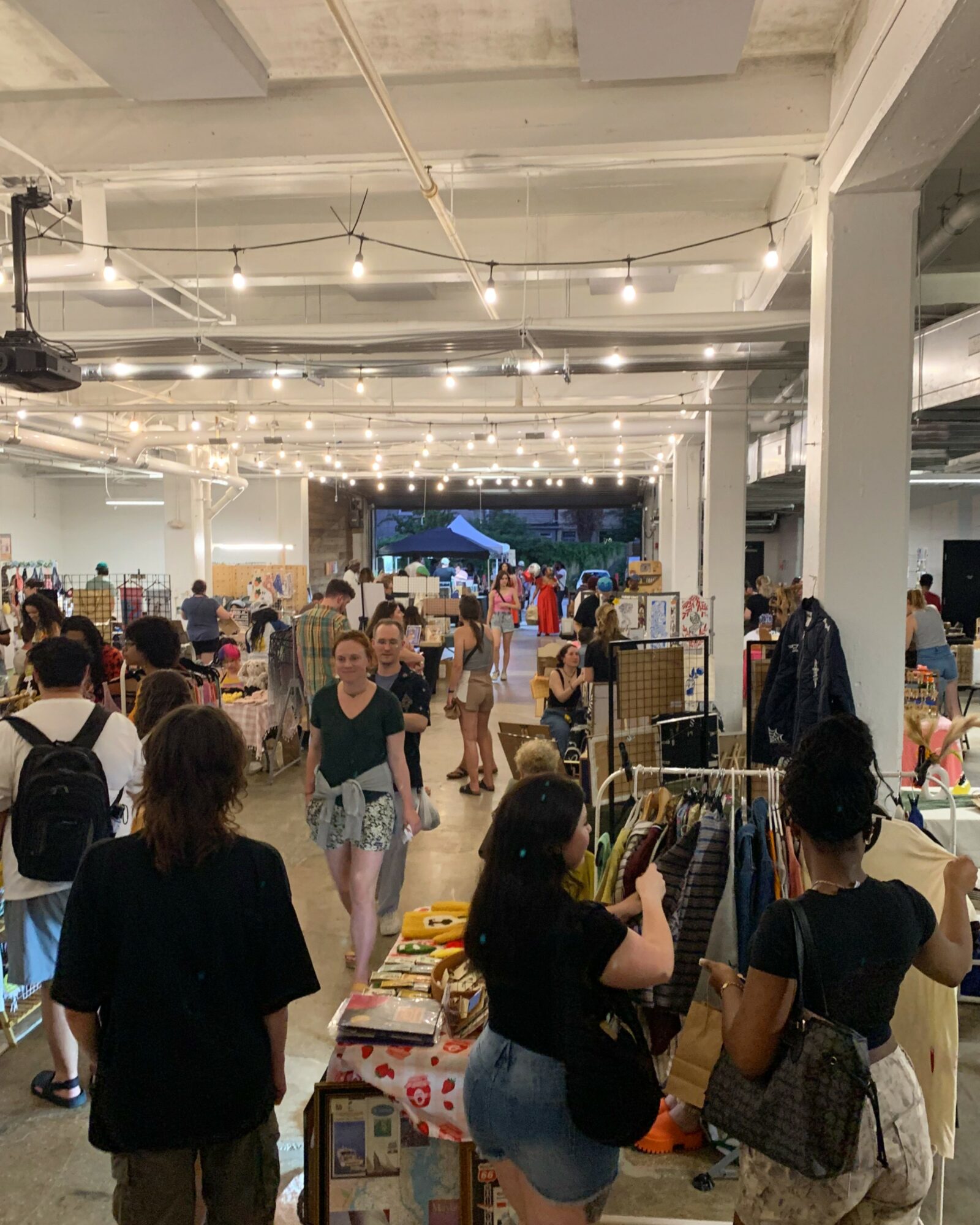 Indoor market with people browsing stalls and hanging string lights, high ceiling, and open space.
