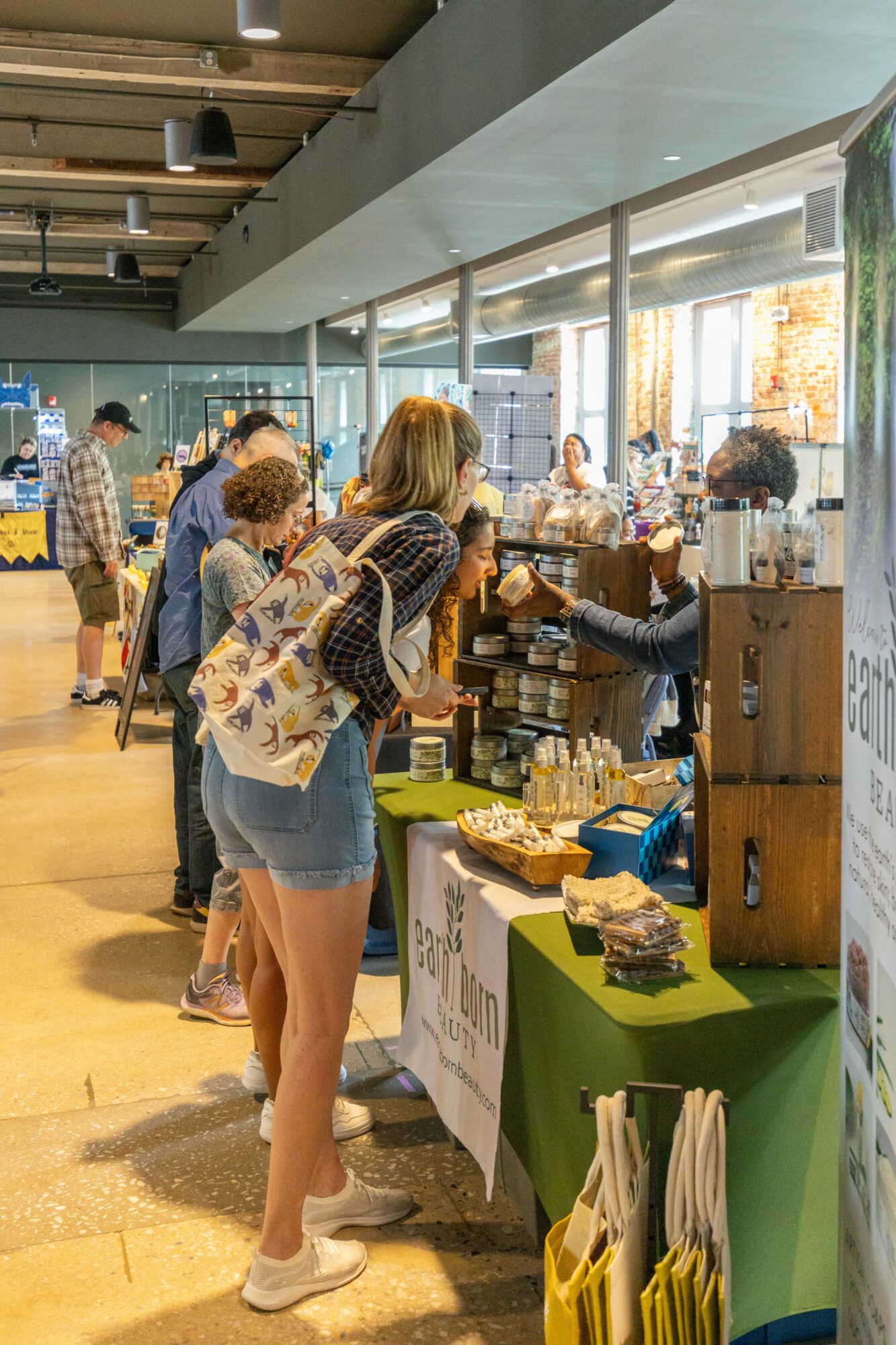 People browsing at a market stall with various products, inside a well-lit indoor space with large windows.