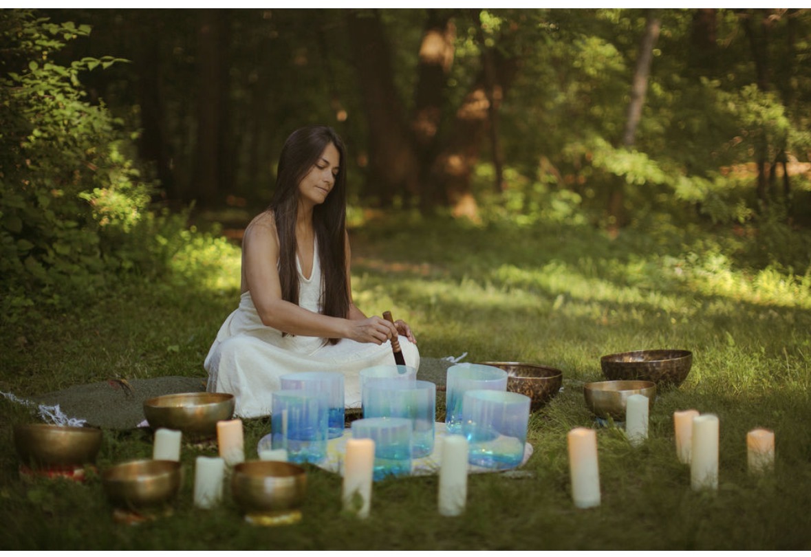 Woman in white dress sitting on grass surrounded by candles and glass containers outdoors, trees in background.