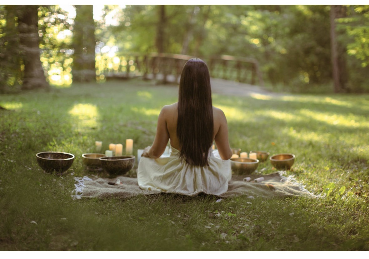 Person with long hair sitting cross-legged on a blanket outdoors, surrounded by bowls and candles, in a forest setting.