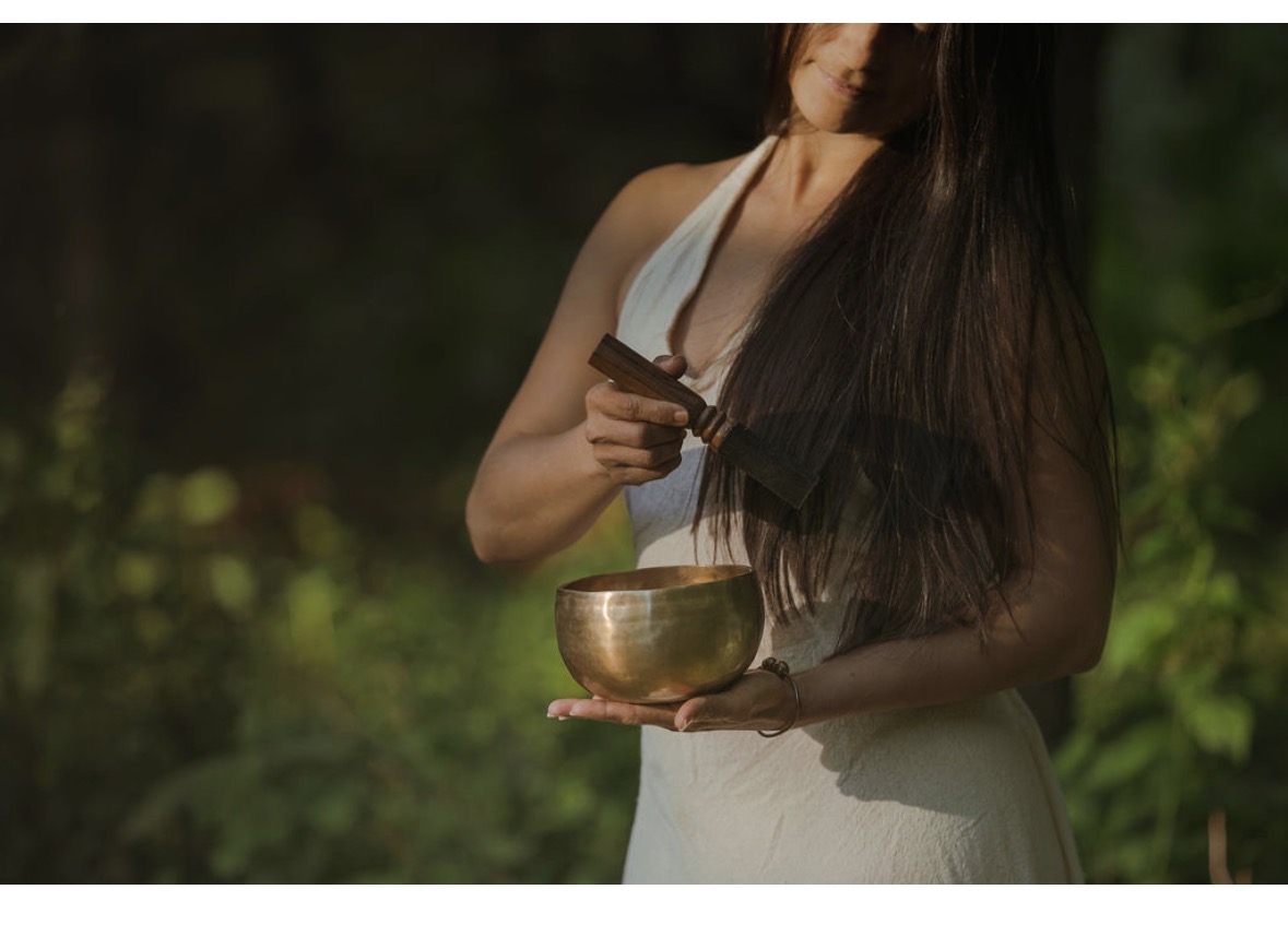 Woman with long dark hair holding a singing bowl outdoors, wearing a white sleeveless dress, surrounded by greenery.
