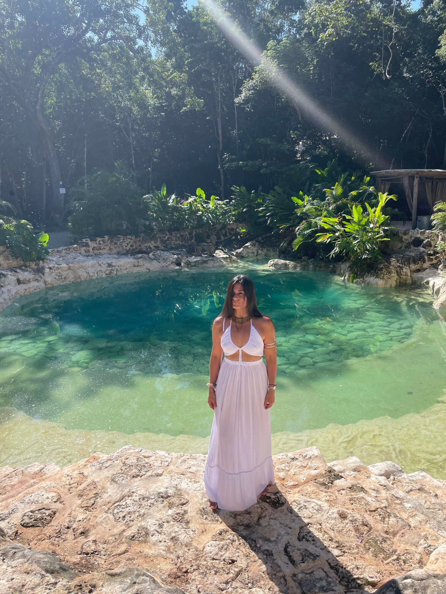 Woman in white dress standing on rocky shore near clear water, surrounded by trees and sunlight.