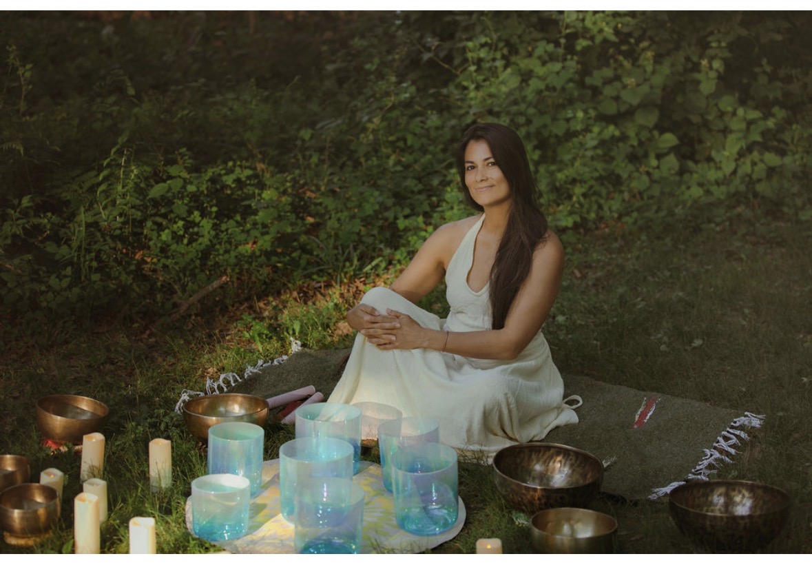Woman in white dress sitting on mat outdoors surrounded by candles and bowls, greenery in background.