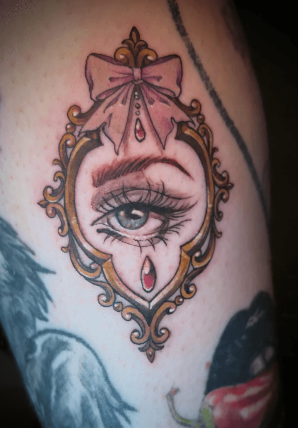 Tattoo of a detailed eye within an ornate frame, decorated with a pink bow and jewel-like accents.