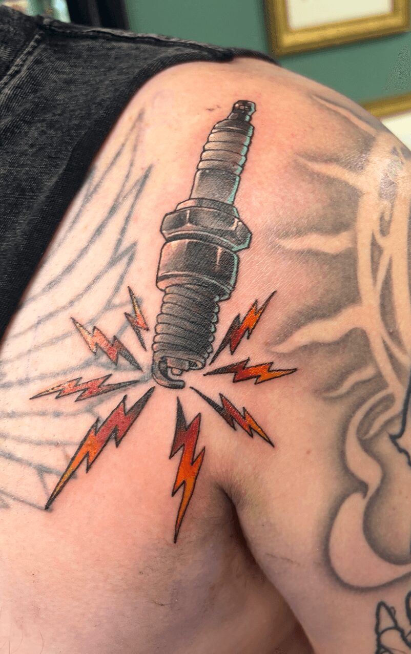 Spark plug tattoo with lightning bolts and sparks on person's shoulder, with part of face visible in background.