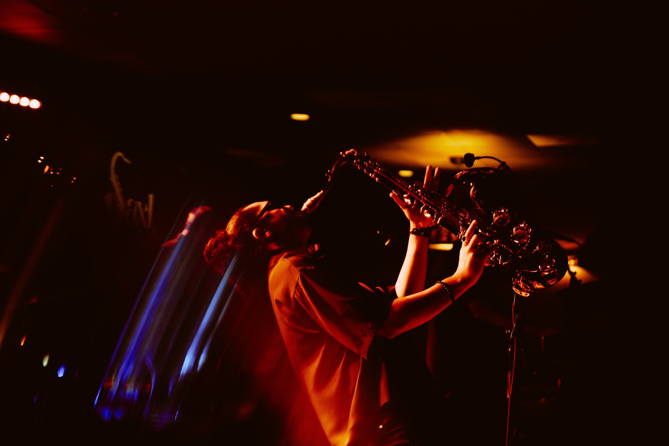 Person playing saxophone on stage with dark background and warm lighting, silhouette visible, holding instrument with both hands.