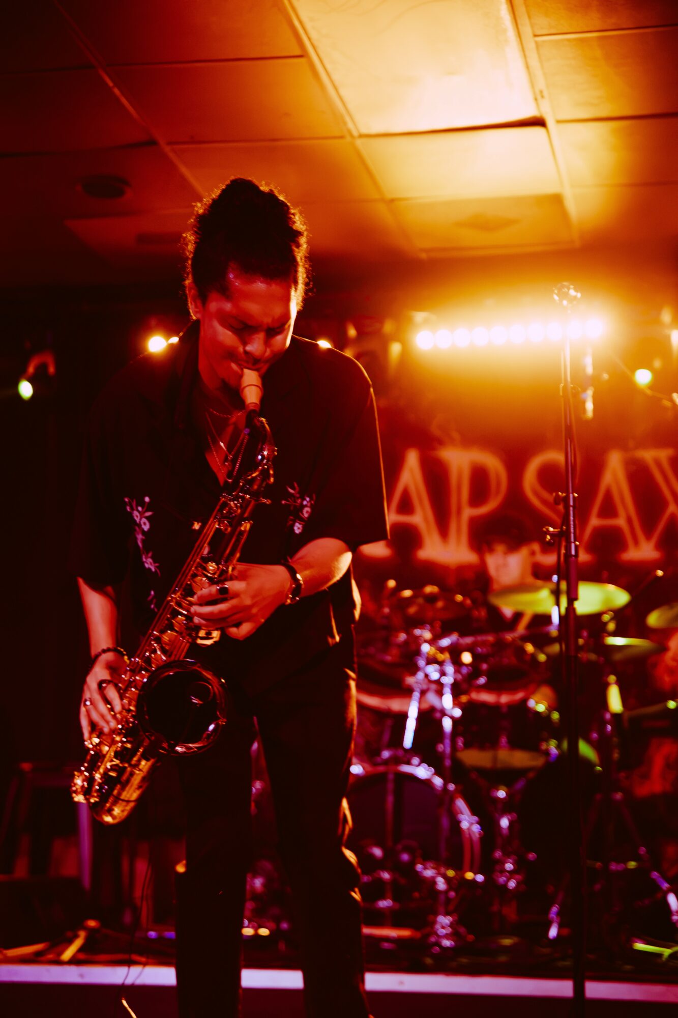 Person playing saxophone on stage with bright lights and drum set in background.