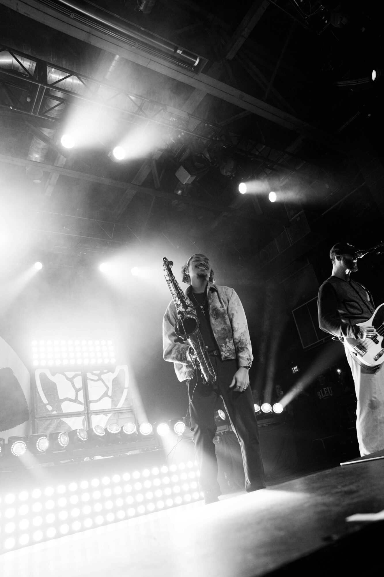Musician with a saxophone performing on stage with bright lights and a guitarist nearby, in black and white.