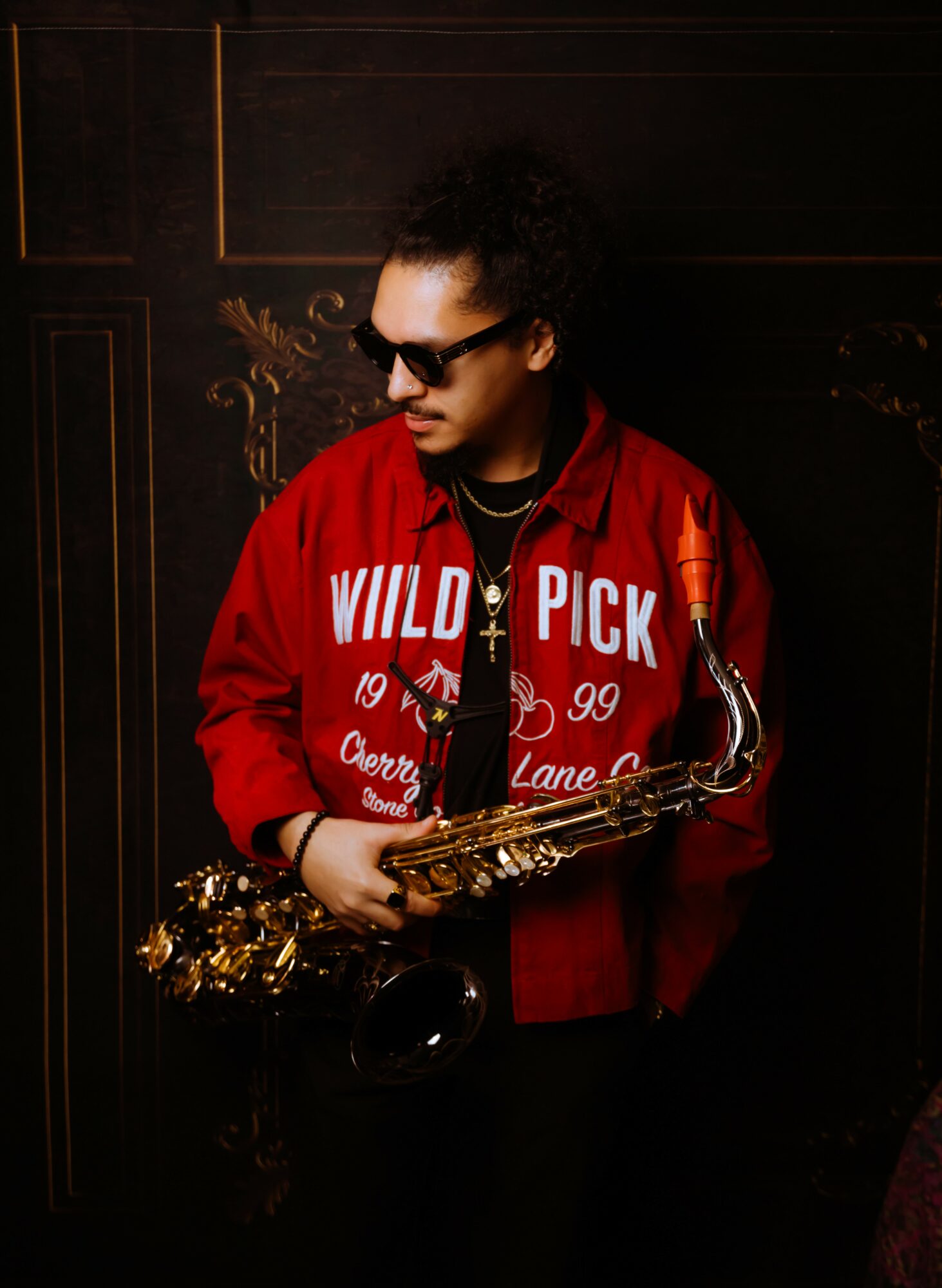 Person with curly hair wearing sunglasses and a red jacket holding a saxophone, standing against a dark background.