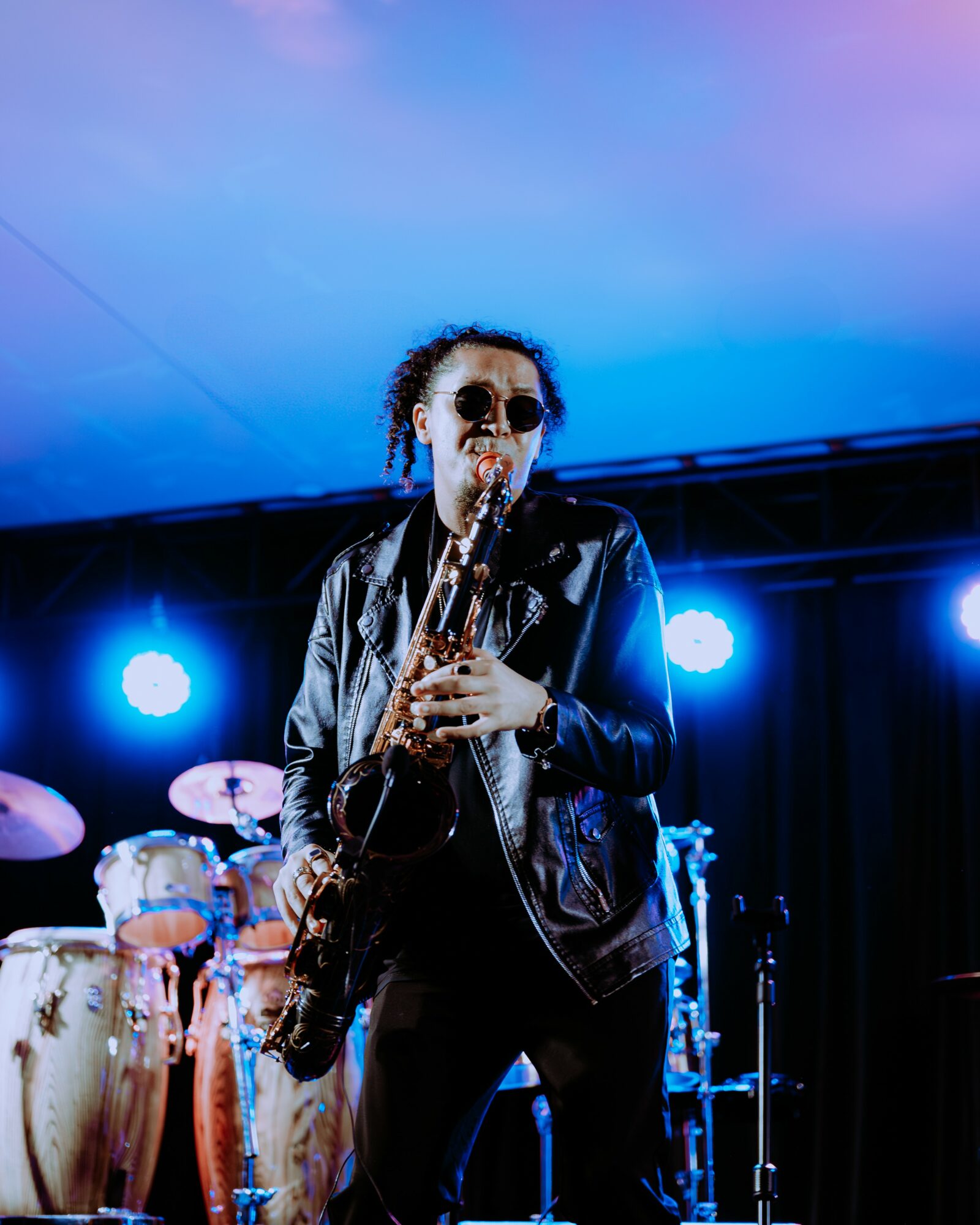Person playing saxophone on stage with blue lighting and musical instruments in background.