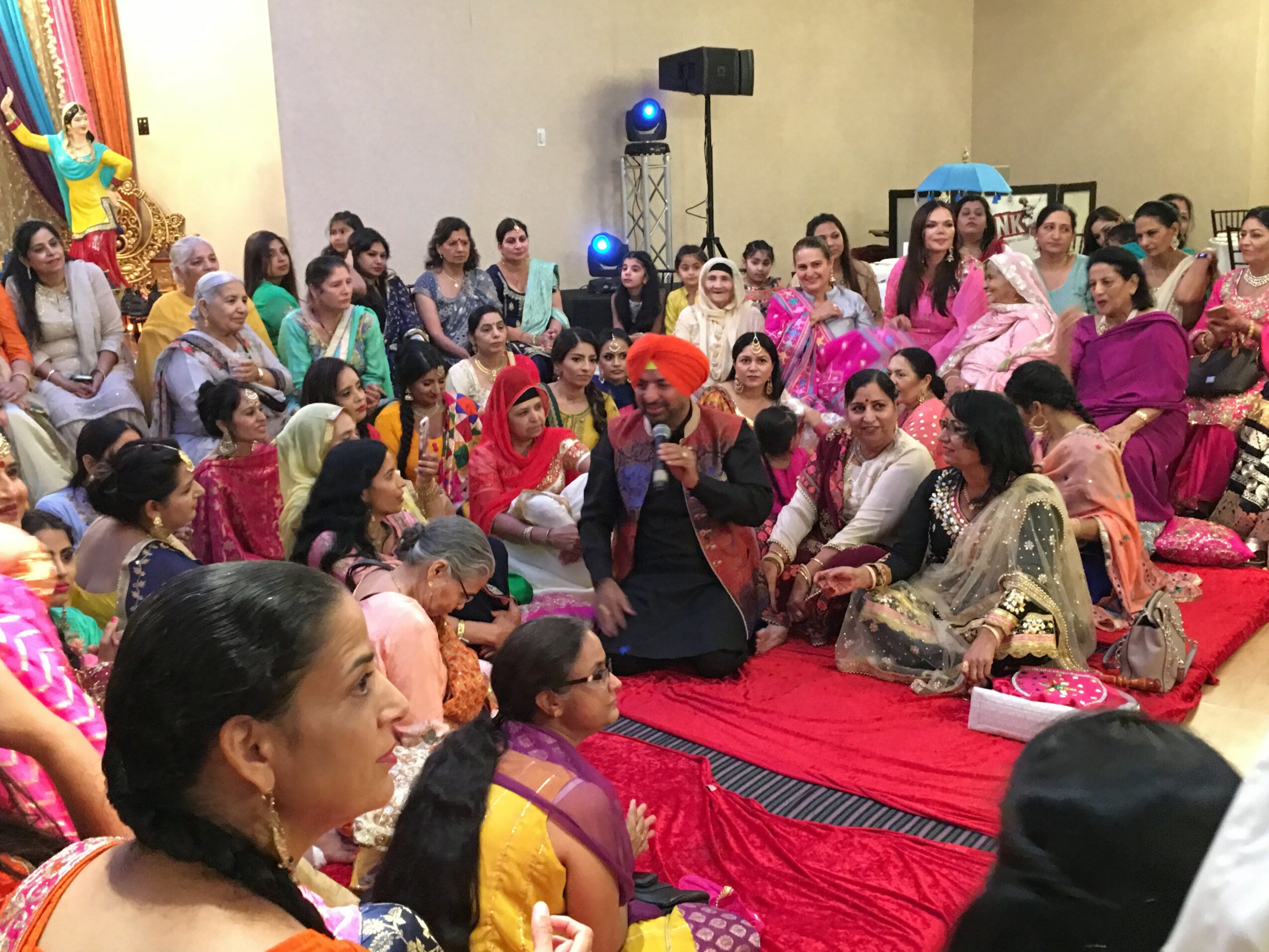 Group of people gathered indoors, sitting on a red carpet, dressed in colorful traditional clothing, attending a cultural event.