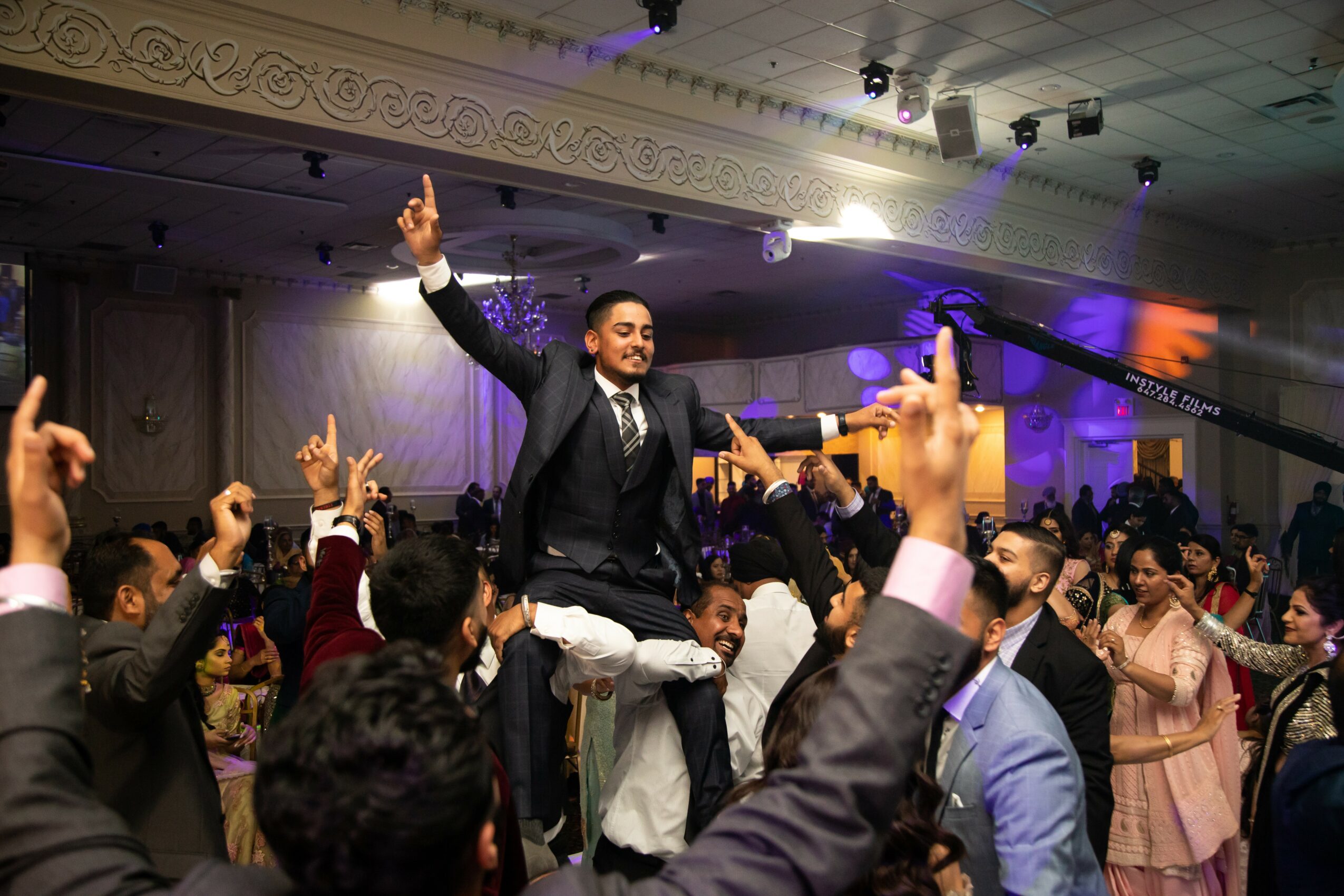 Man in a suit is lifted by people at a celebration, with others raising hands and cheering in a decorated hall.