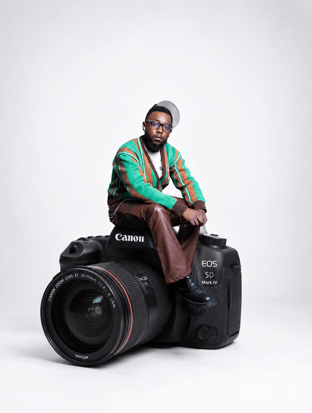 Person sitting on a large Canon EOS 5D camera with a plain white background.