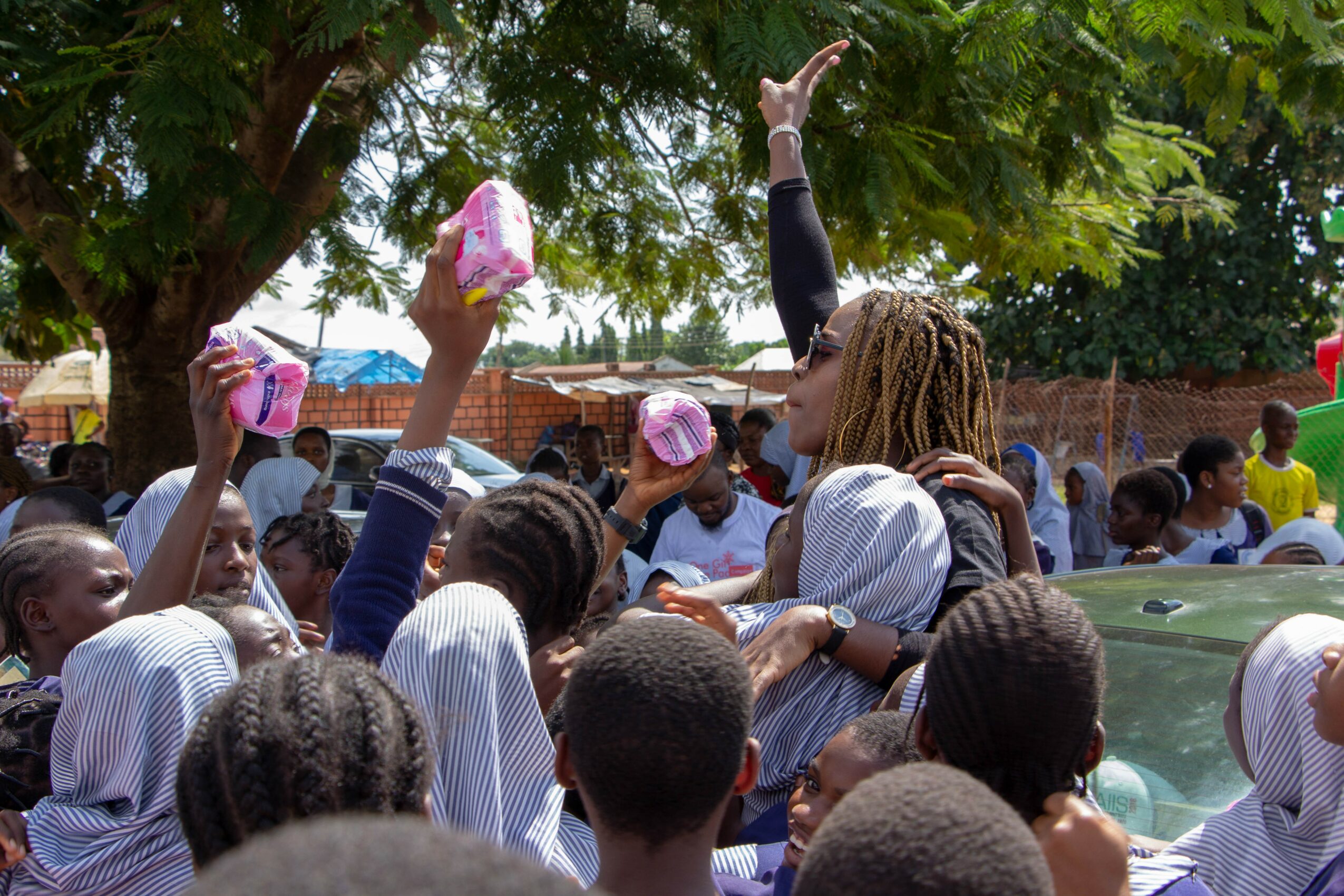 A woman with dreadlocks interacts with children outdoors, some holding pink items, under a tree with a crowd around.