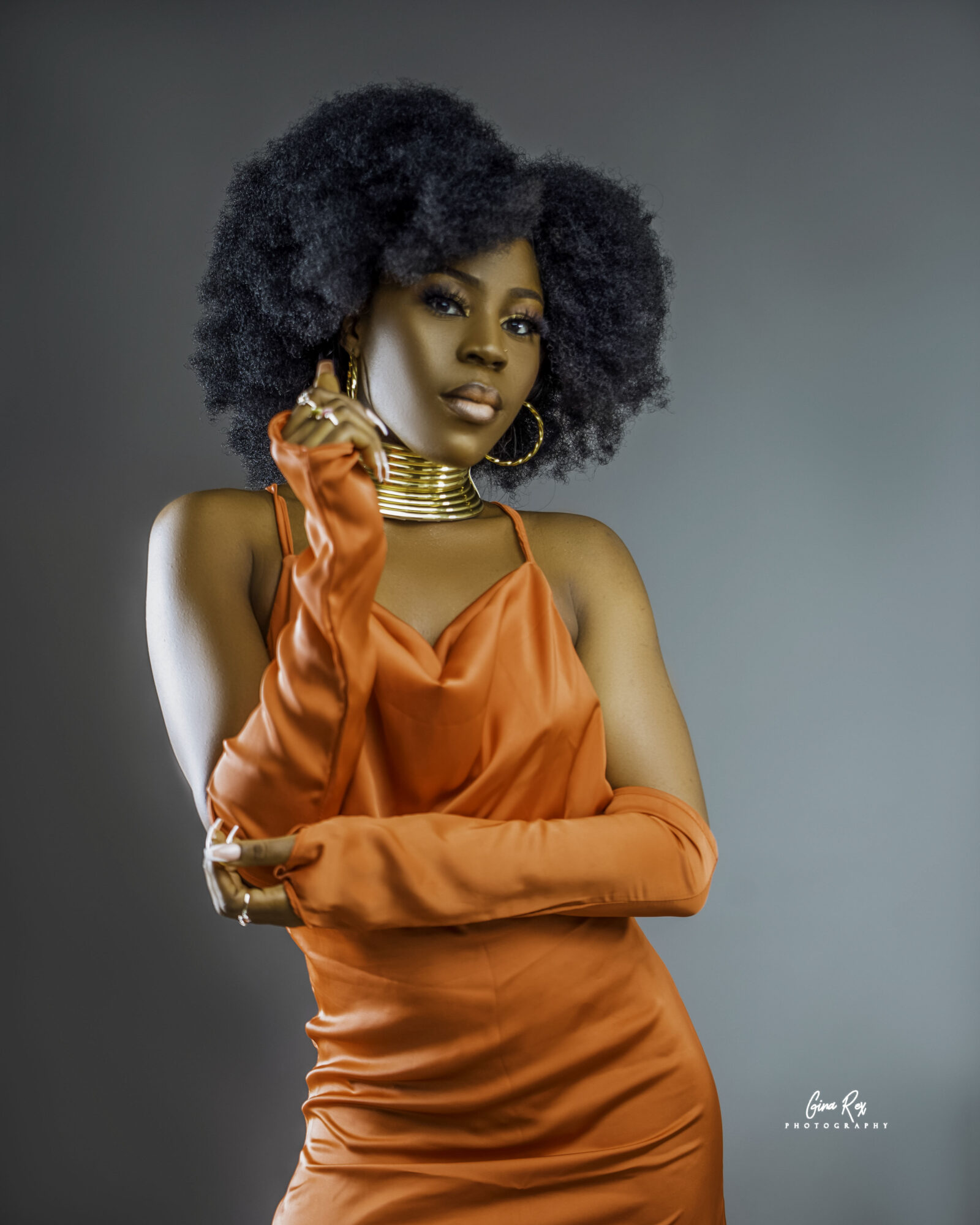 Woman with natural curly hair wearing an orange dress and long glove, posing with hand near face, against a gray background.
