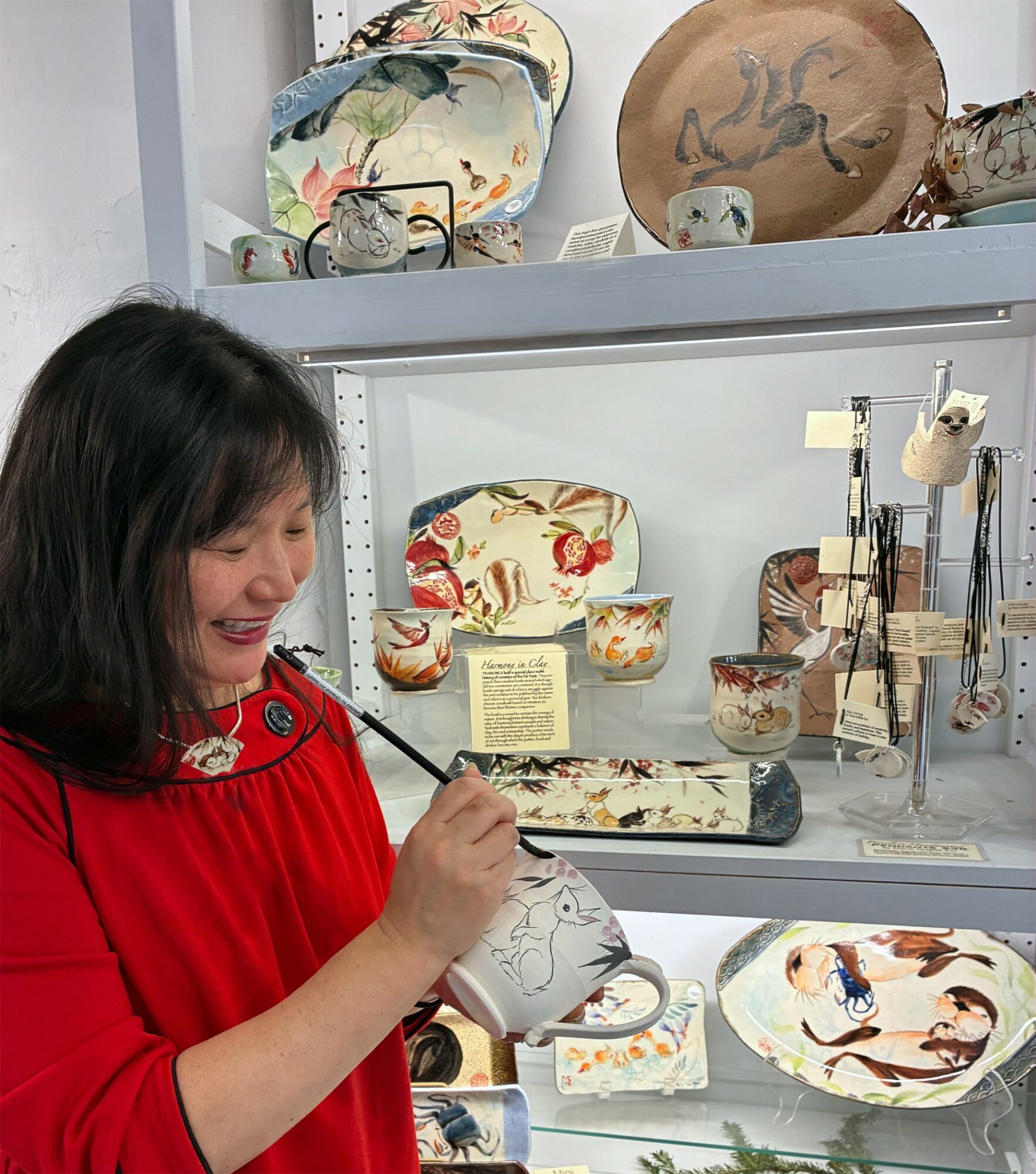 Woman in red dress smiling while painting a ceramic bowl, display of pottery and ceramics behind her.