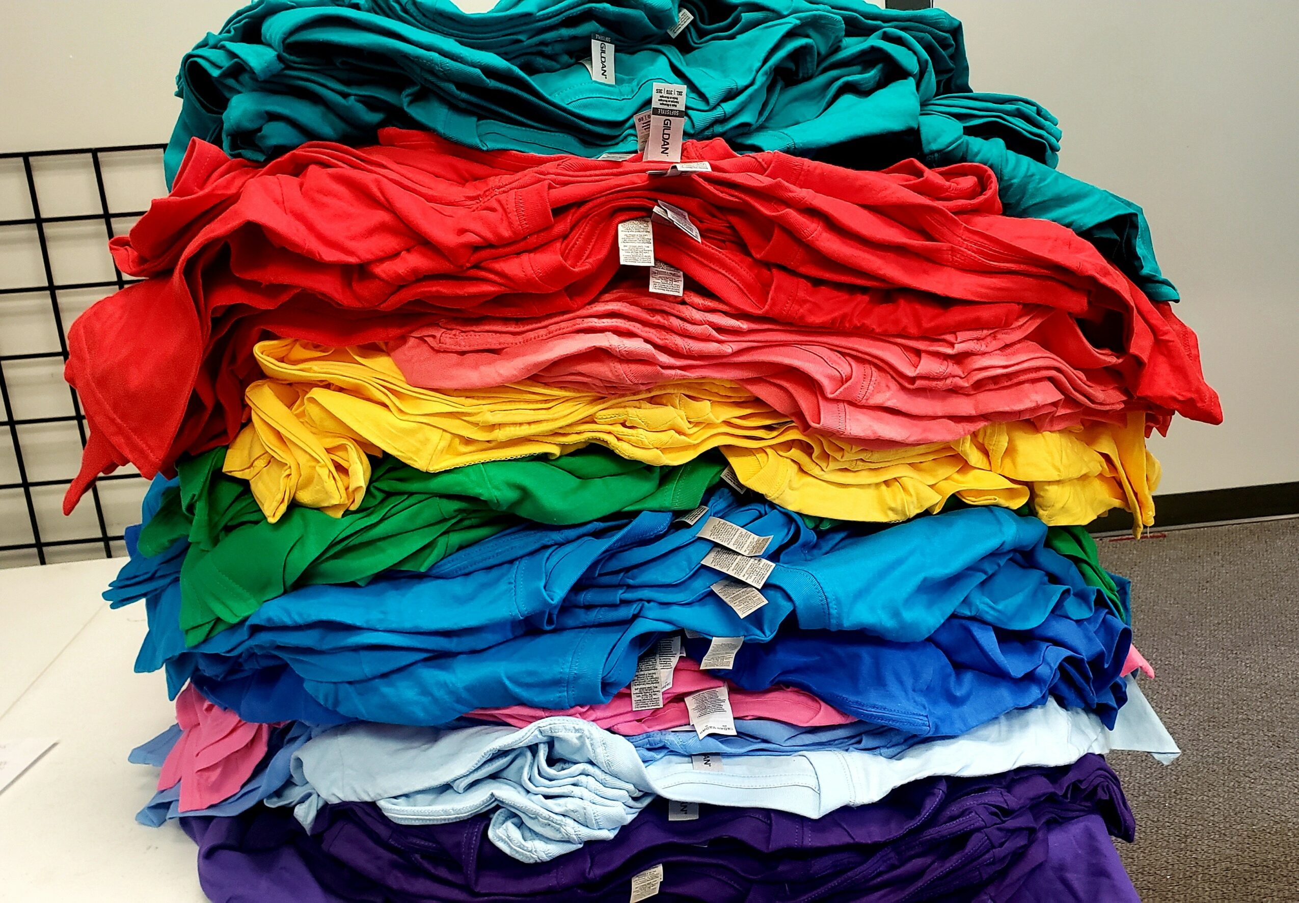 Stack of colorful folded T-shirts in teal, red, yellow, green, blue, white, and purple on a table.
