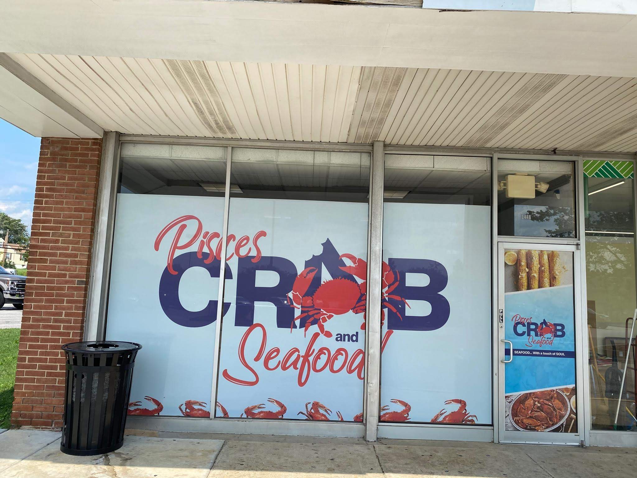 Storefront with large window displaying a seafood restaurant sign, crab illustrations, and a door with a poster, trash can outside.