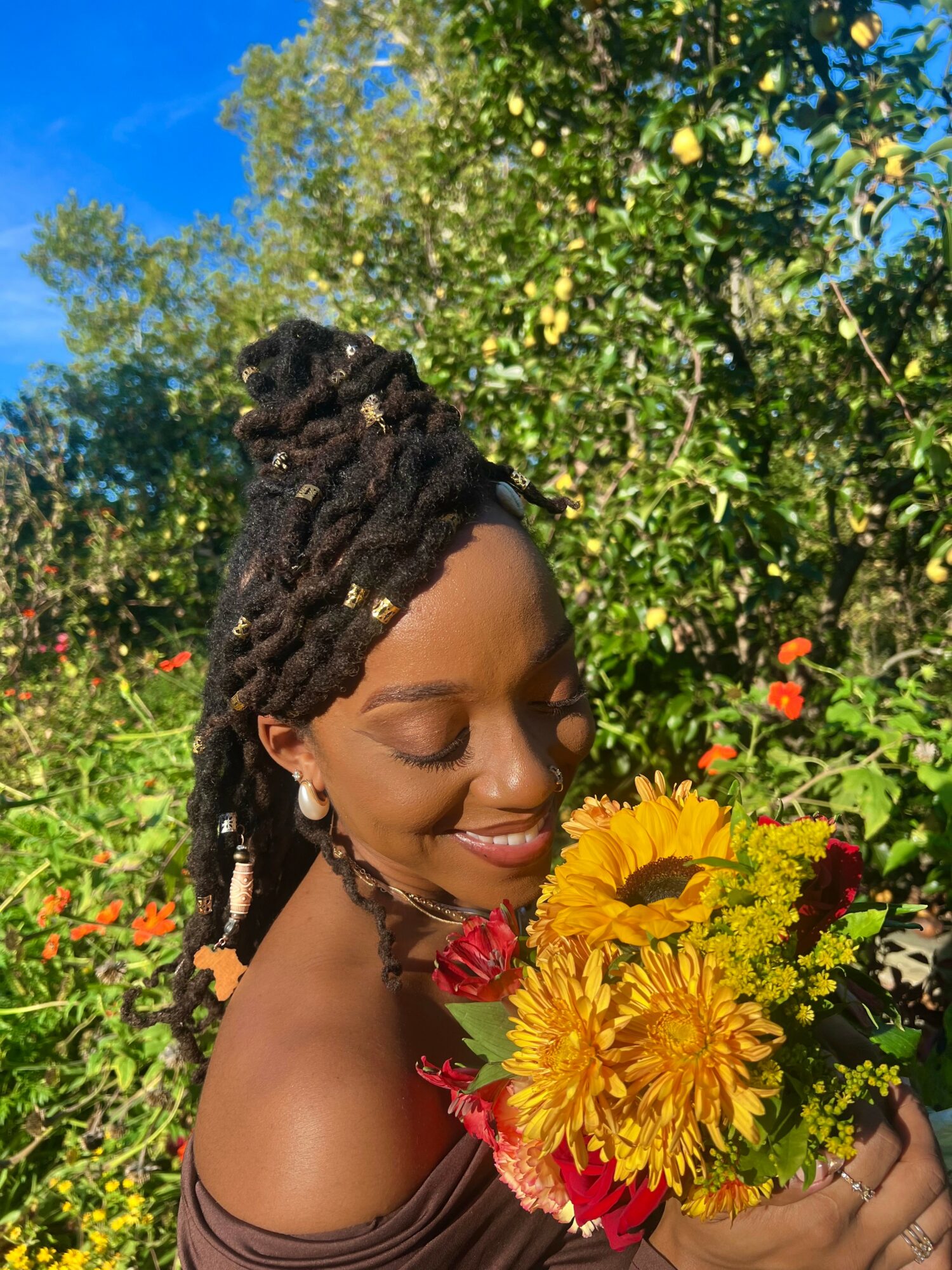 Smiling woman with dreadlocks holding a colorful bouquet of flowers outdoors, surrounded by green trees and blue sky.